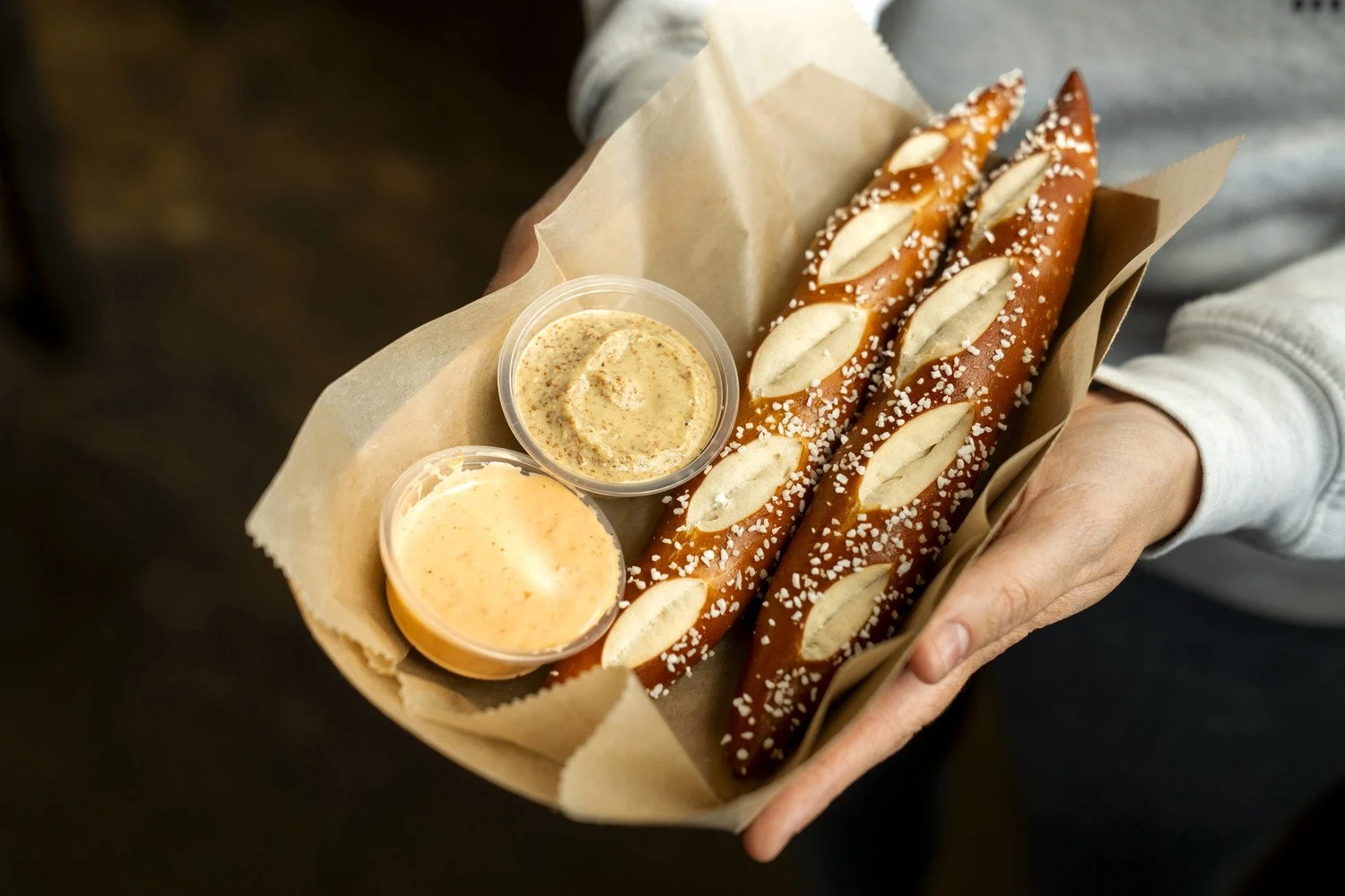Person holding a tray with two long pretzels sprinkled with coarse salt, served with two dipping sauces