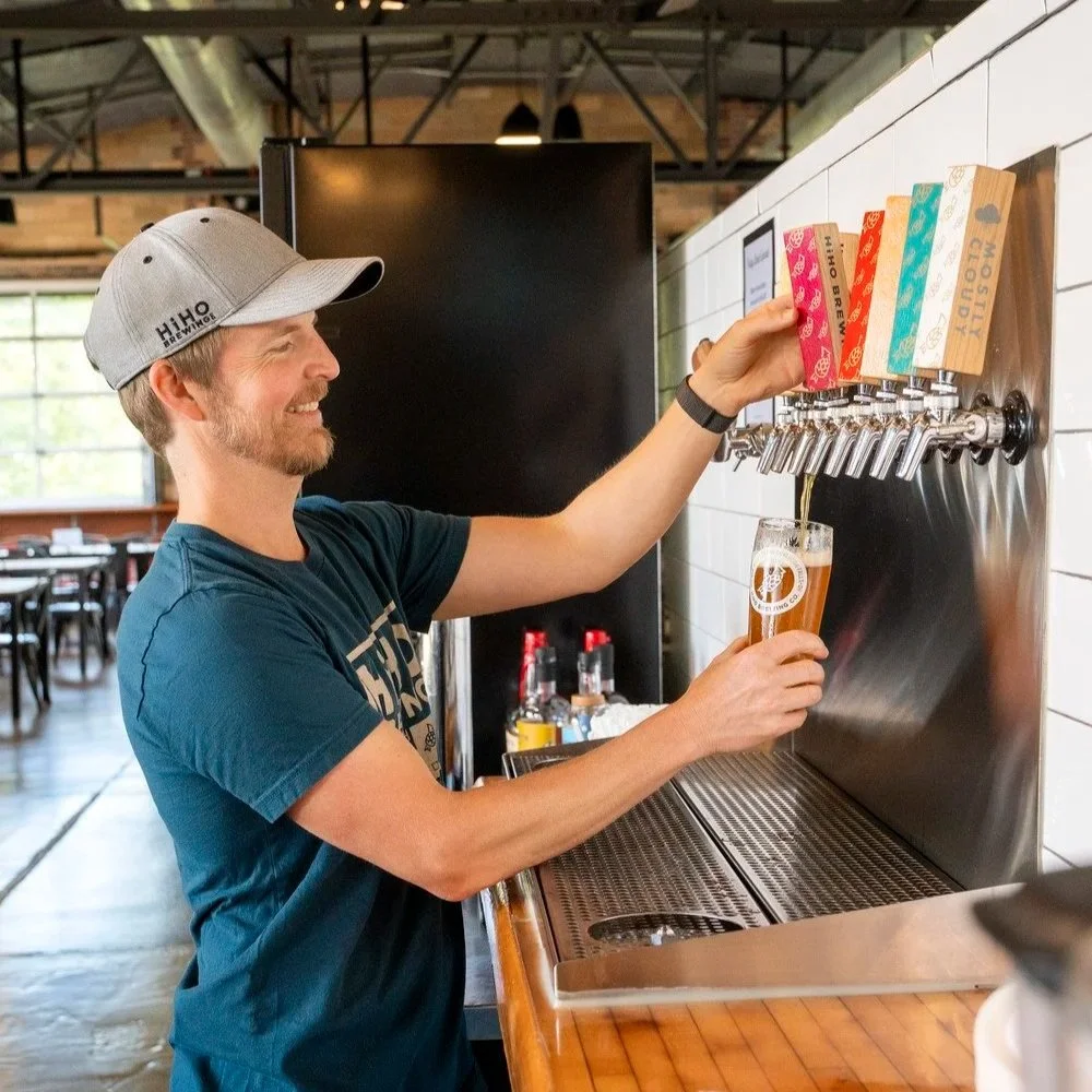 A man wearing a gray baseball cap and a blue T-shirt pours a glass of beer from a tap at a HiHO Brewing Co.