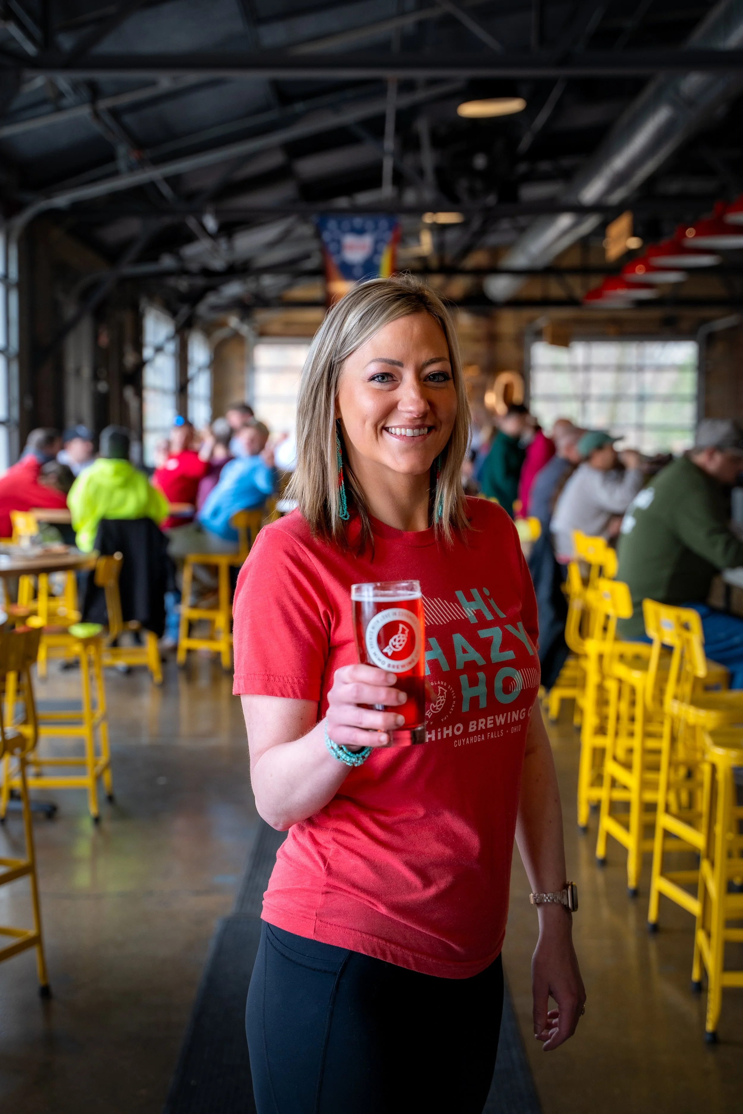 A woman smiling and holding a glass of beer in a lively brewery or bar with many people seated at tables and yellow chairs in the background.