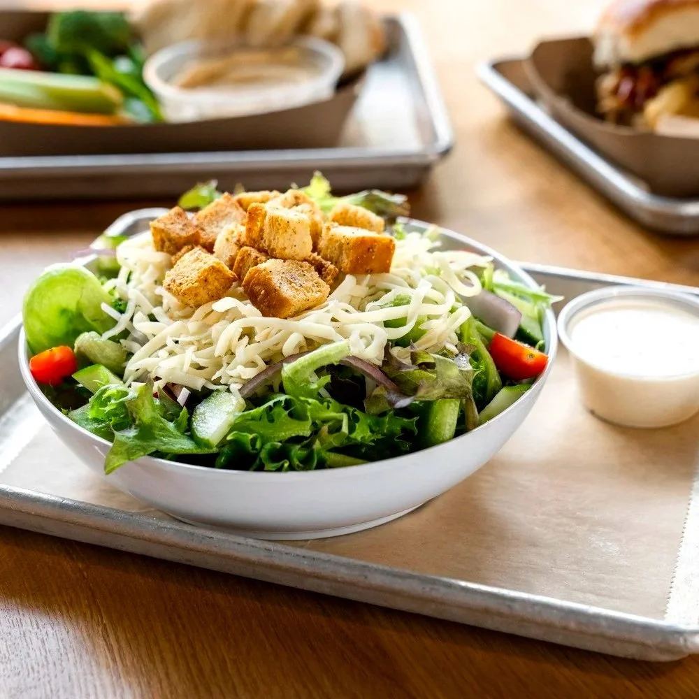 Small house salad with croutons, shredded cheese, cherry tomatoes, cucumbers, and leafy greens, served on a tray with a side of salad dressing.