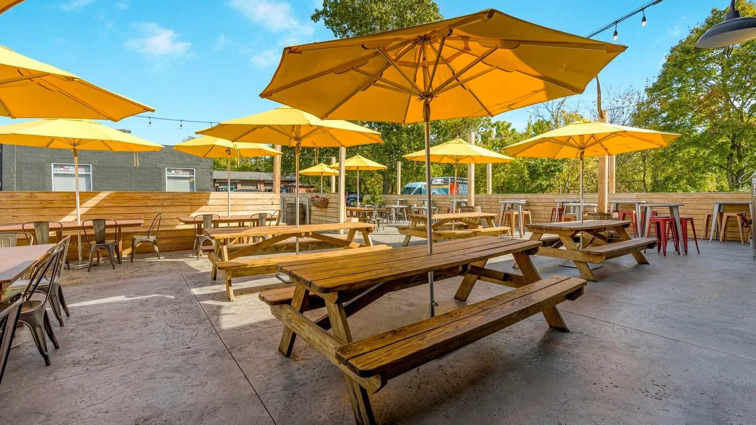 Outdoor patio area with wooden picnic tables and yellow umbrellas under a blue sky with some trees in the background.