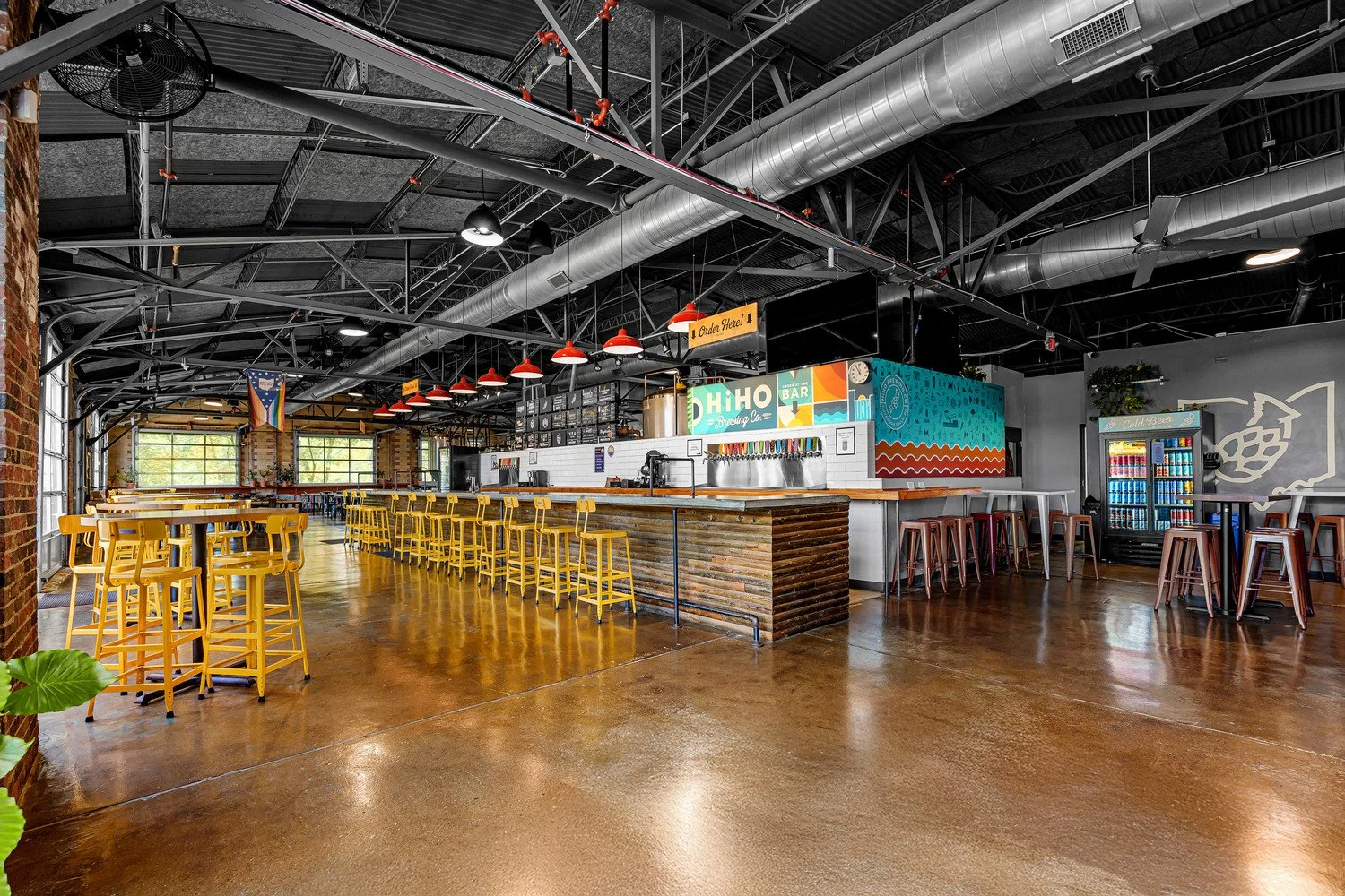 Interior of HiHO Brewing Co. with yellow chairs, a bar with colorful artwork, and a refrigerator full of cans of 4-packs. The space features an industrial style with exposed ductwork, black ceiling, and large windows letting in natural light.