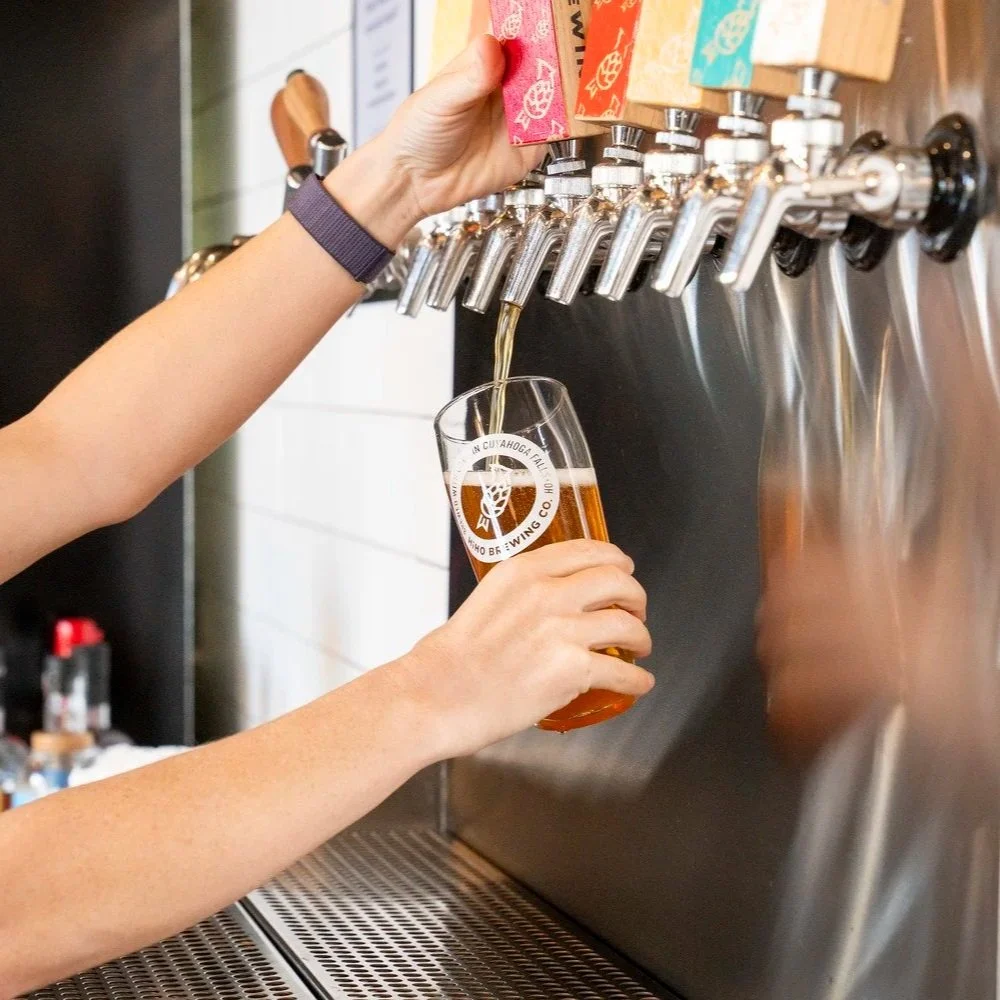 A person is pouring beer from a tap into a glass, inside HiHO Brewing Co. The tap system has 20 beer taps and colorful labels.