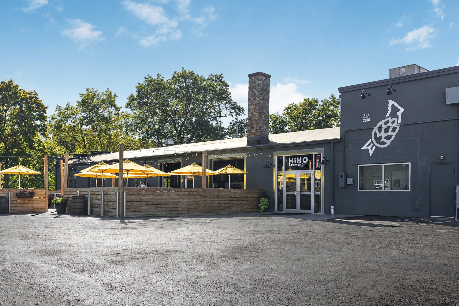 Exterior view of a modern building with outdoor seating, yellow umbrellas, and a painted logo of a hop cone and bird on the wall, indicating it is a brewery named HiHO Brewing Co., established in 2016.