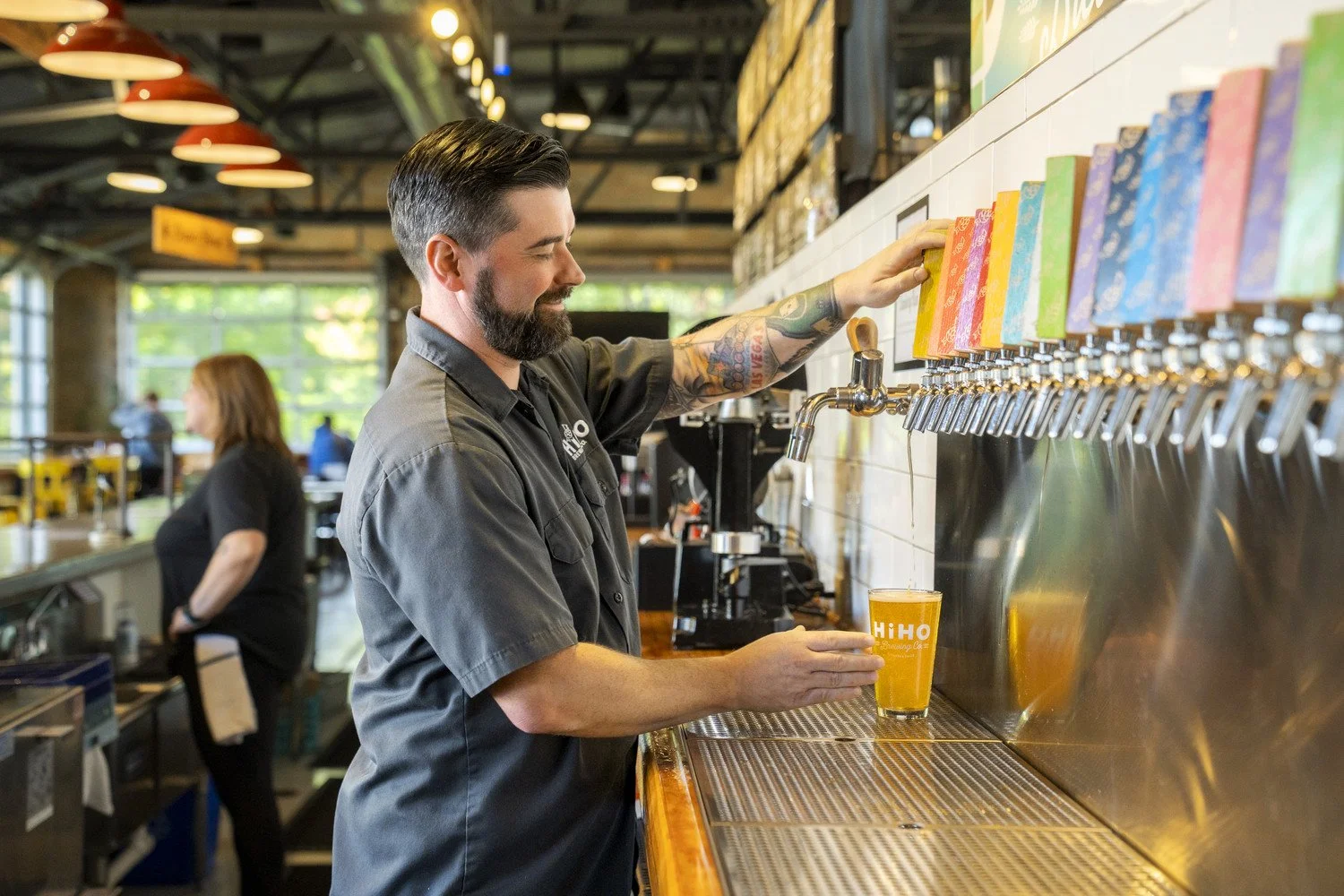 A bartender with a beard and tattoos pours beer from a tap into a glass at a bar with colorful beer taps.