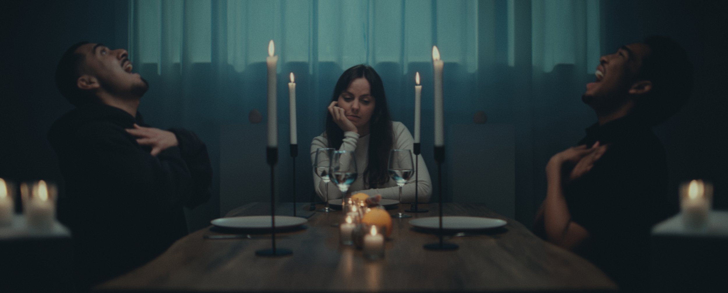 Three people sit at a dimly lit dining table illuminated by candles. The two men on either side are laughing loudly with their heads thrown back, while the woman sitting between them rests her chin on her hand, looking unamused.