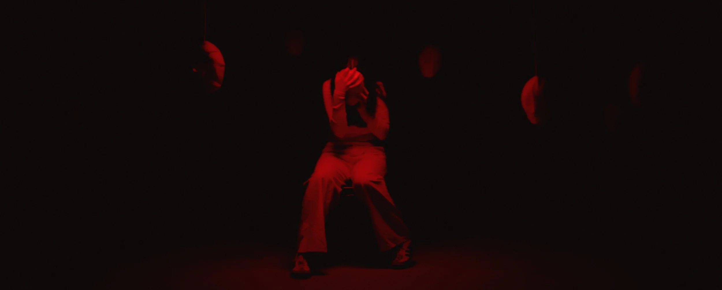 A person sits hunched over, holding their head in a dark room illuminated by intense red light, surrounded by hanging, featureless white masks.