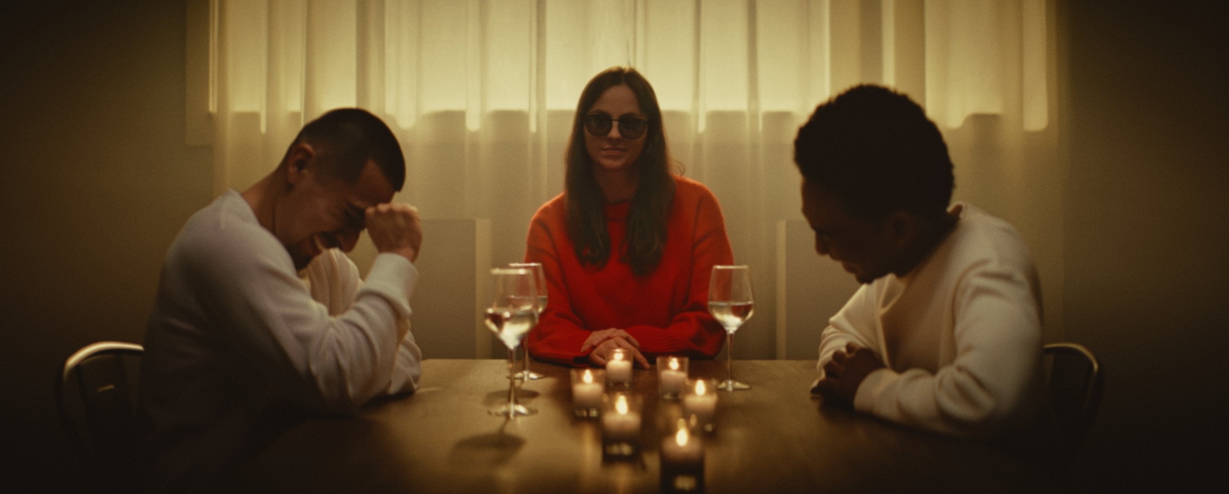 A moody, cinematic shot of a smiling woman wearing a red sweater and dark sunglasses sitting between two men laughing at a dinner table illuminated by small candles.