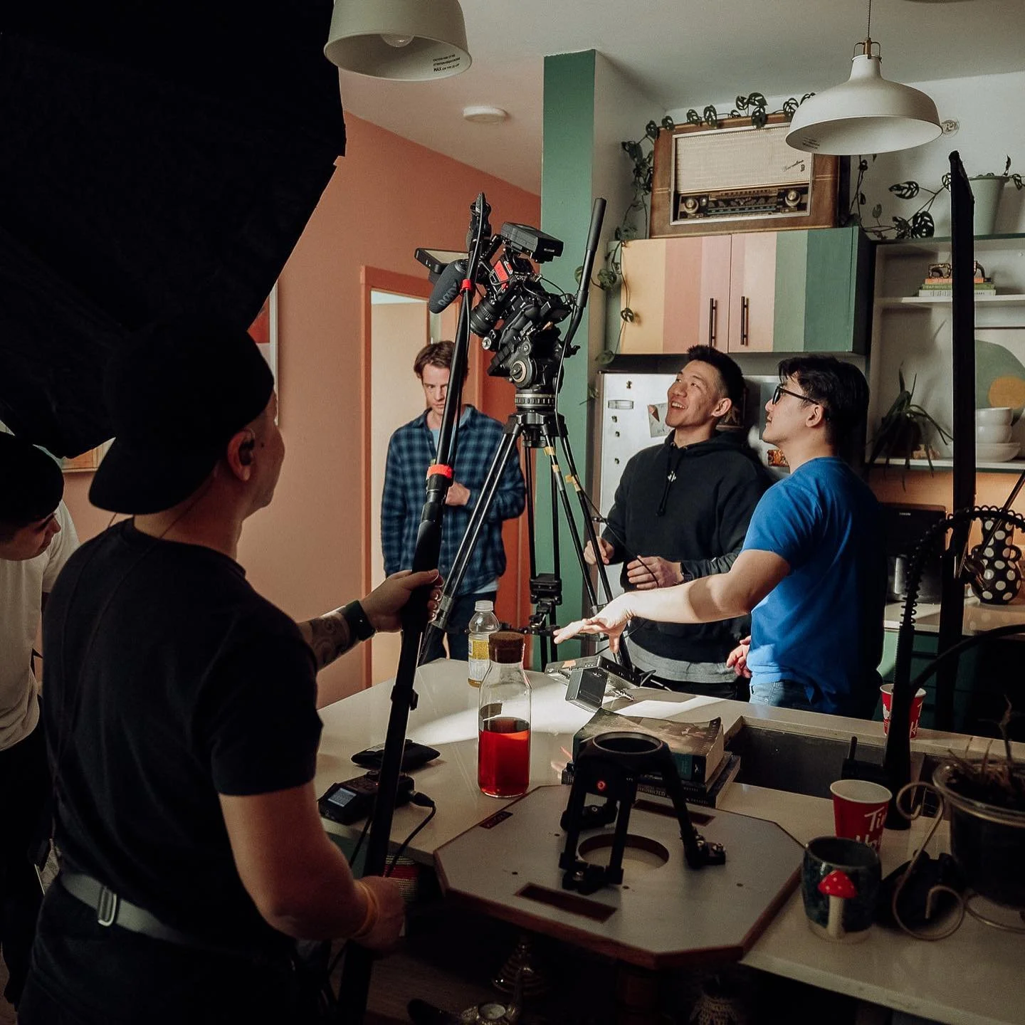 People filming a video in a cozy kitchen with colorful cabinets, plants, and vintage decor.