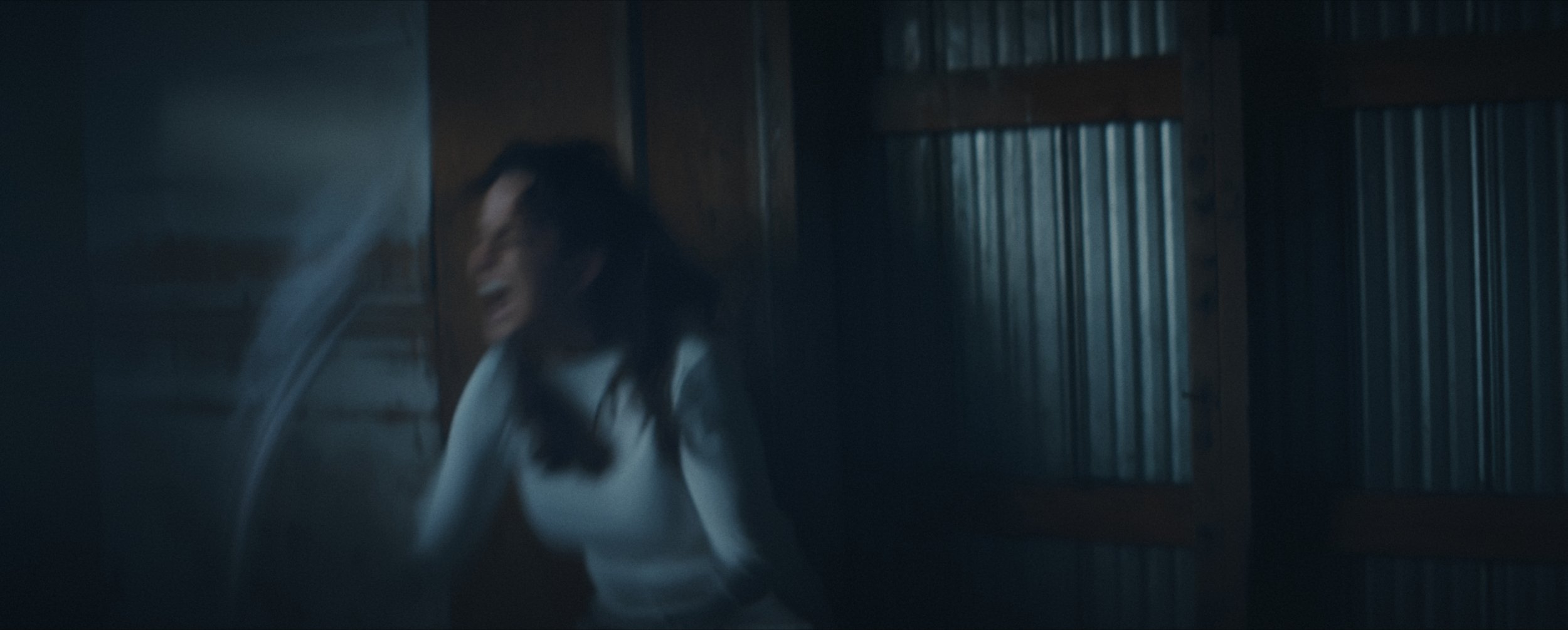 A dark, blurry action shot of a woman in a white shirt shouting in anger while ripping off a mask.