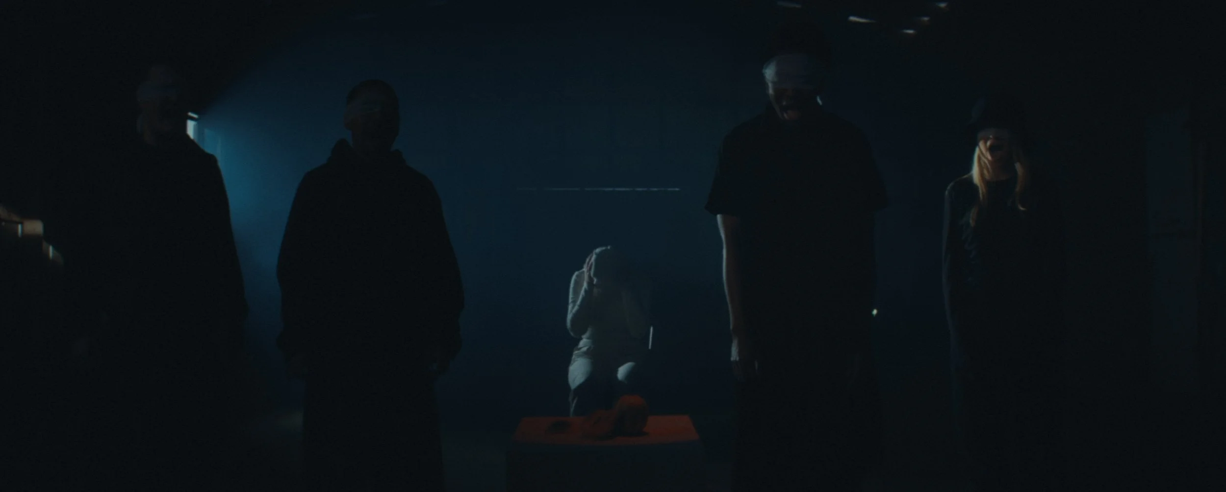 A very dark, blue-lit room where four standing figures wearing dark clothing and white blindfolds surround a distressed person sitting in the background covering their ears.