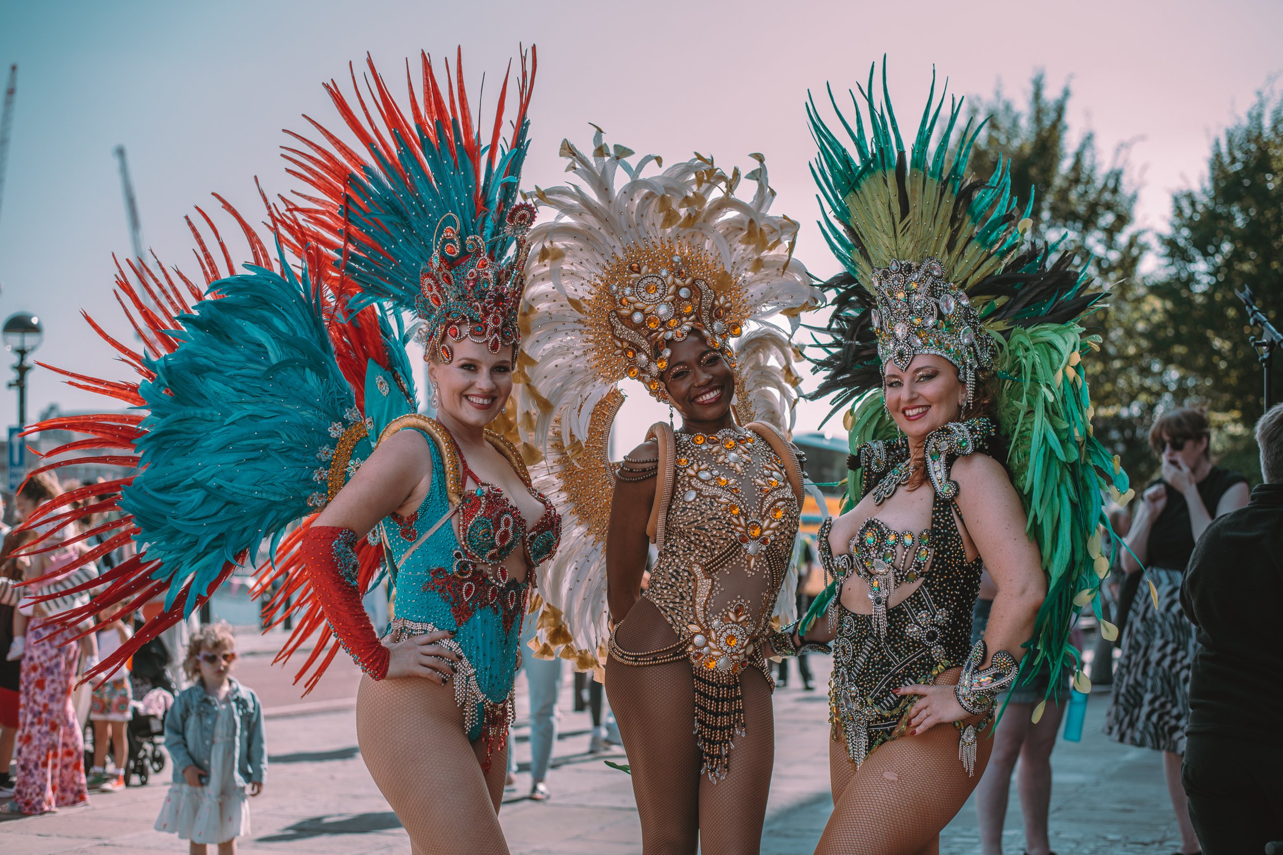 Three women in colorful, elaborate carnival costumes with large feathered headdresses standing outdoors.