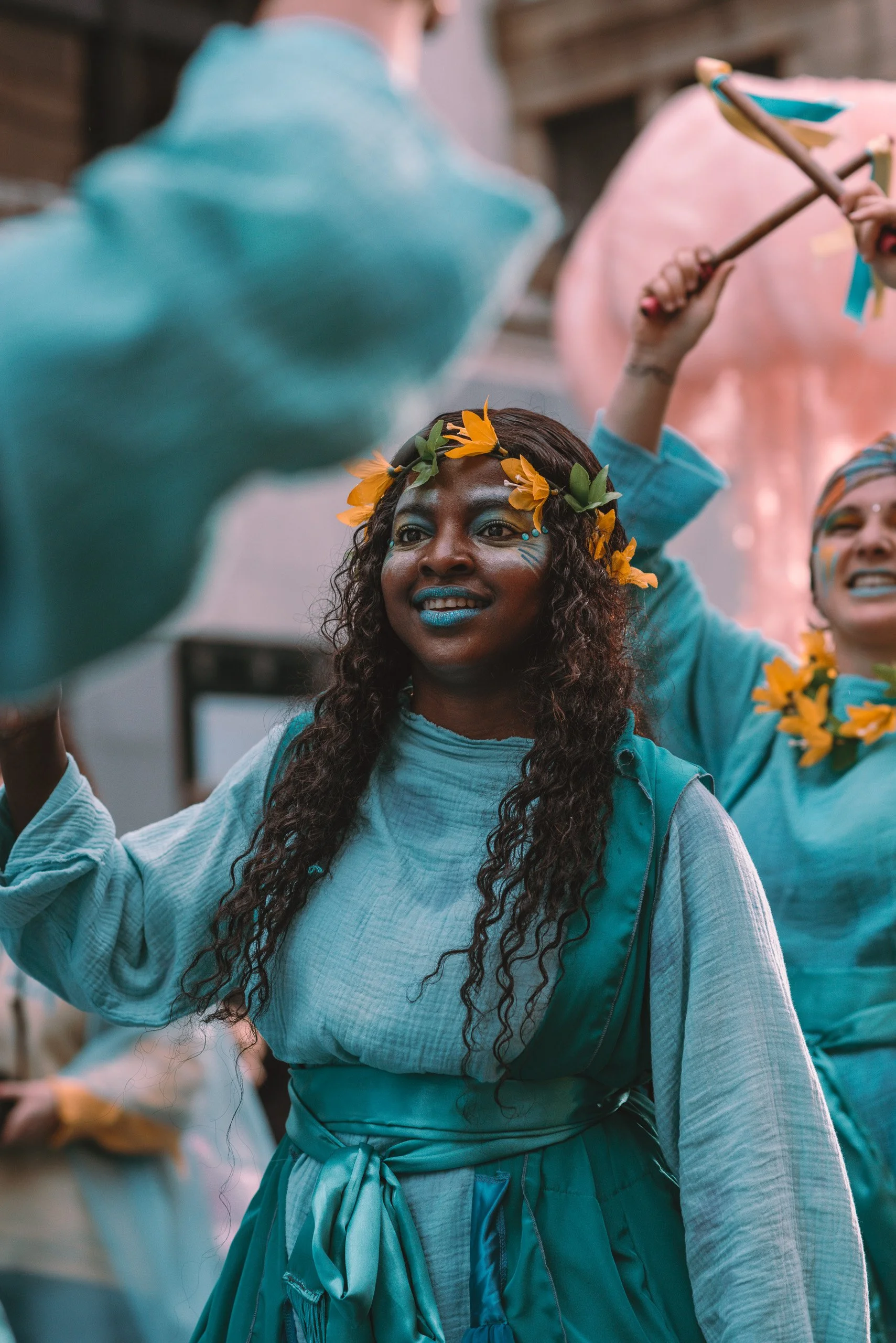 A woman with curly hair wearing a flower crown and face paint, holding a stick with blue and yellow decorations, participating in a celebration or parade.