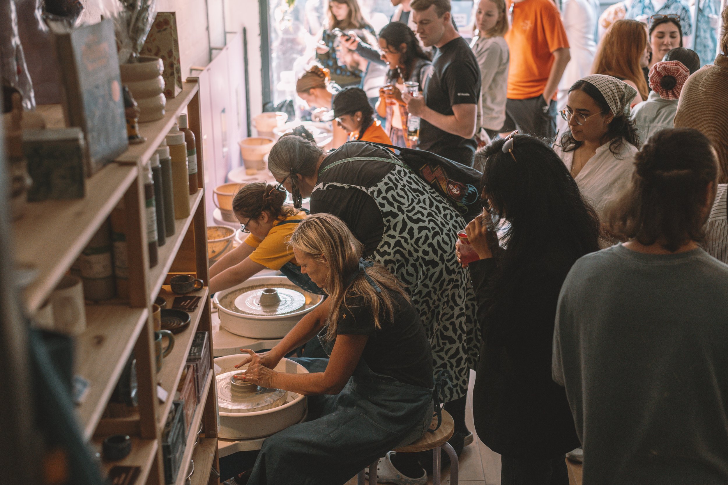 People participating in a pottery class with some working on wheel pottery and others observing, in a cozy studio with shelves holding pottery supplies.