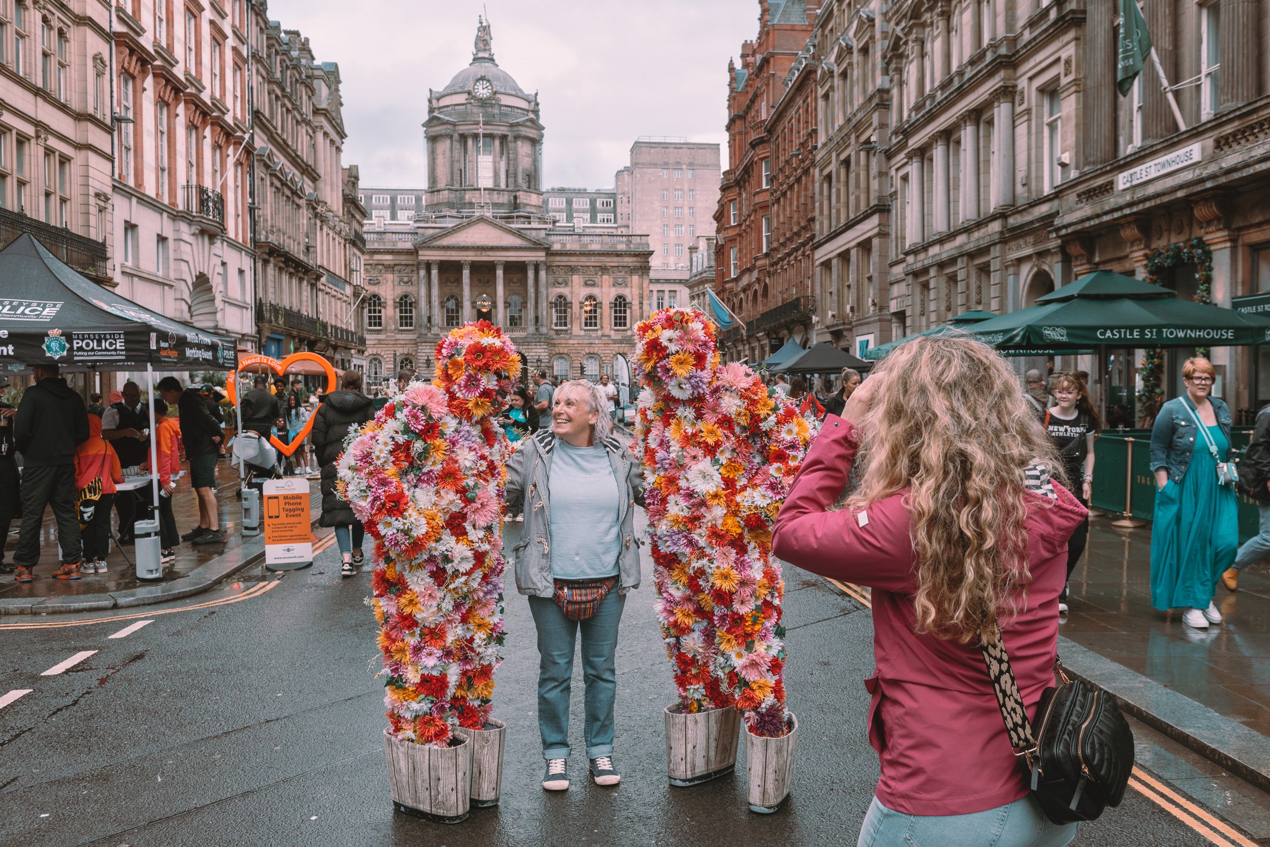 A woman is posing with her face between two large structures covered in pink, orange, and red flowers on a city street, while another woman takes a photo of her. The street is busy with pedestrians and has historic buildings in the background, with u