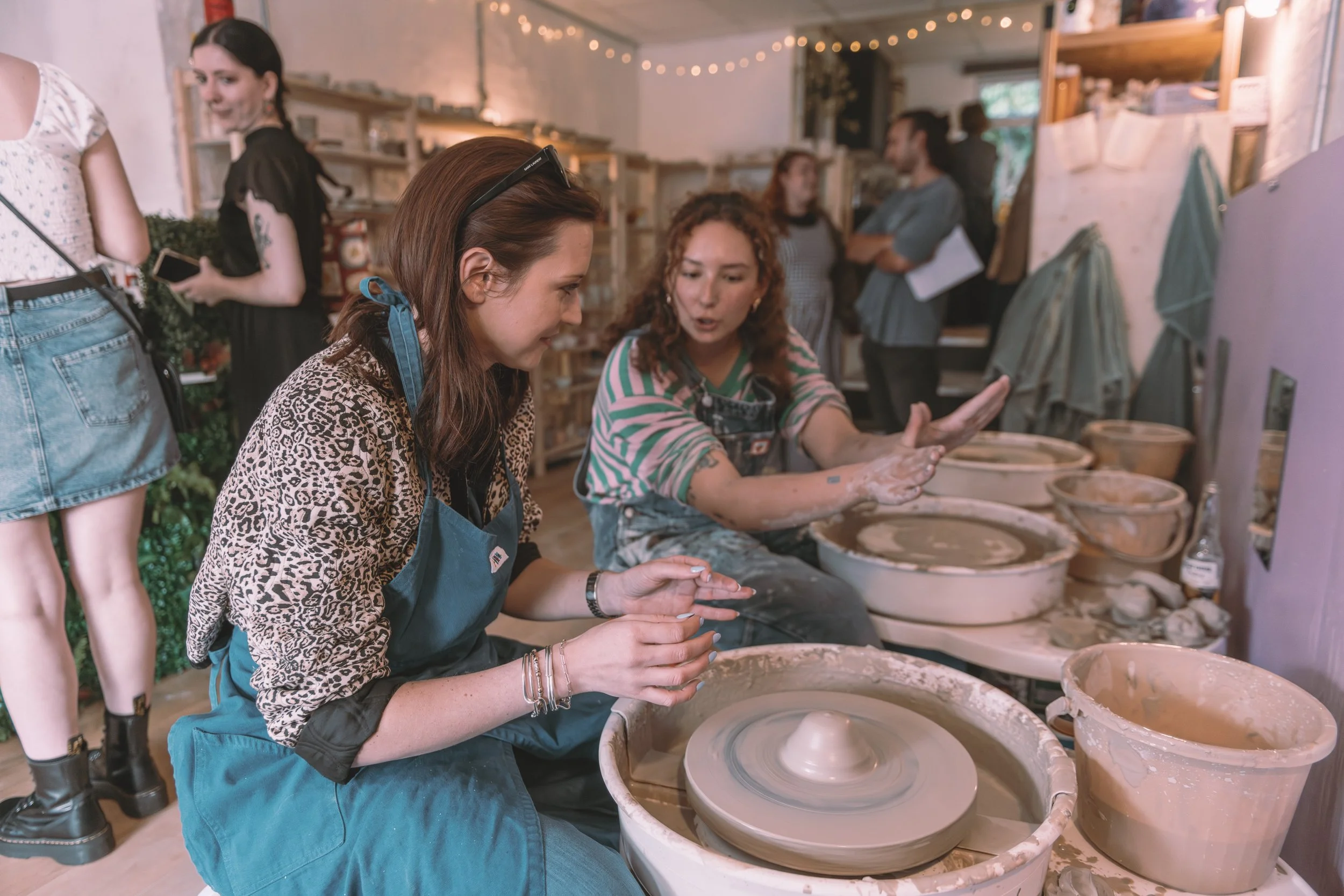 Two women engaging in pottery making at a pottery studio, with pottery wheels and clay in front of them, while others observe in the background.