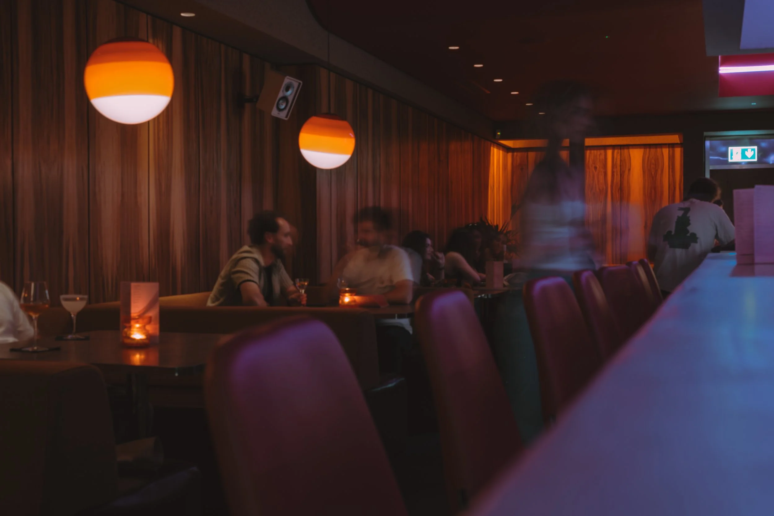 Dimly lit bar or lounge with wooden walls, orange pendant lights, and patrons sitting at tables, some with drinks, with a person standing near the bar.