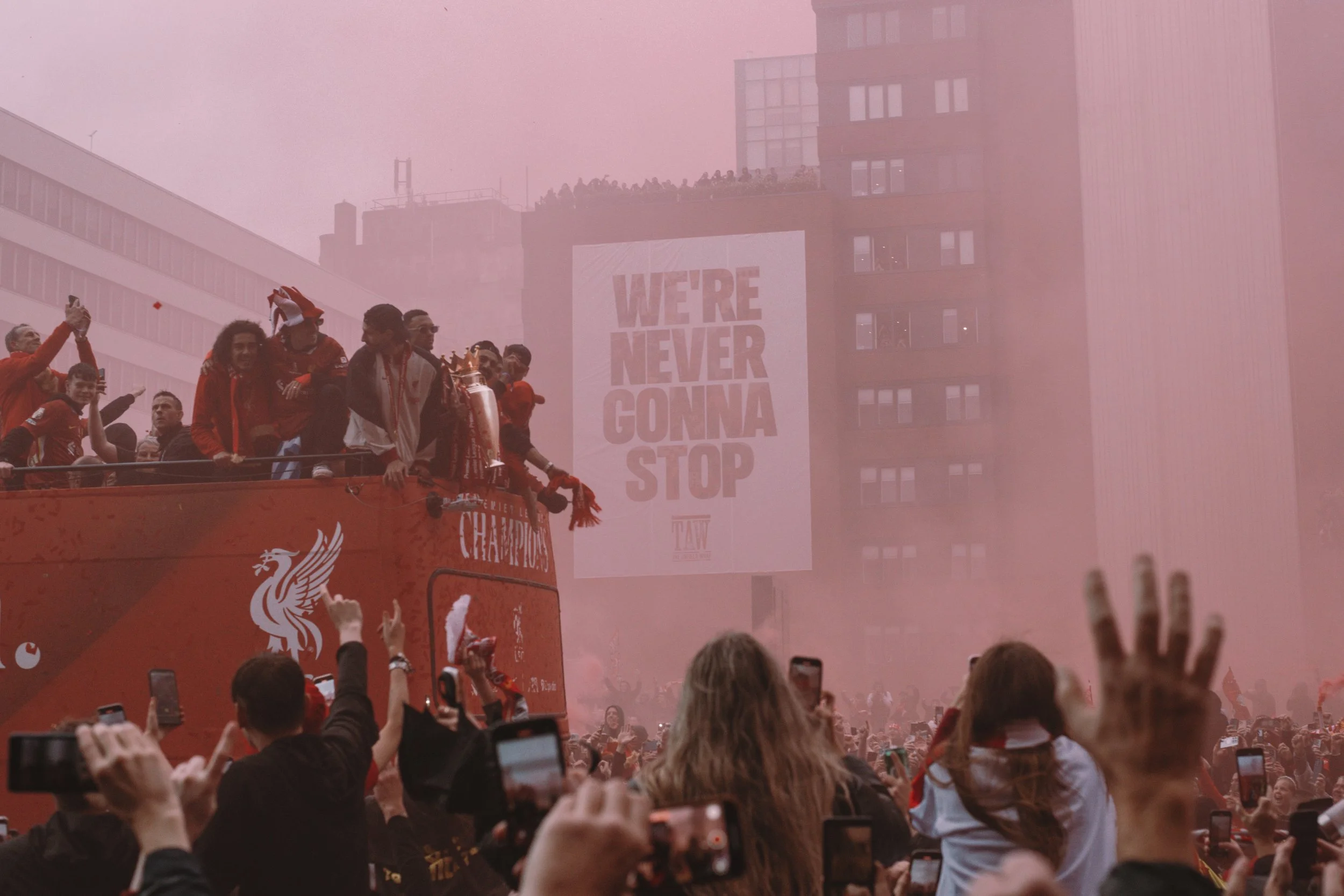 Soccer team celebrating on a parade float with a large crowd in the foreground taking photos. A pink smoke cloud is in the background with a building showing a large sign that reads "WE'RE NEVER GONNA STOP."