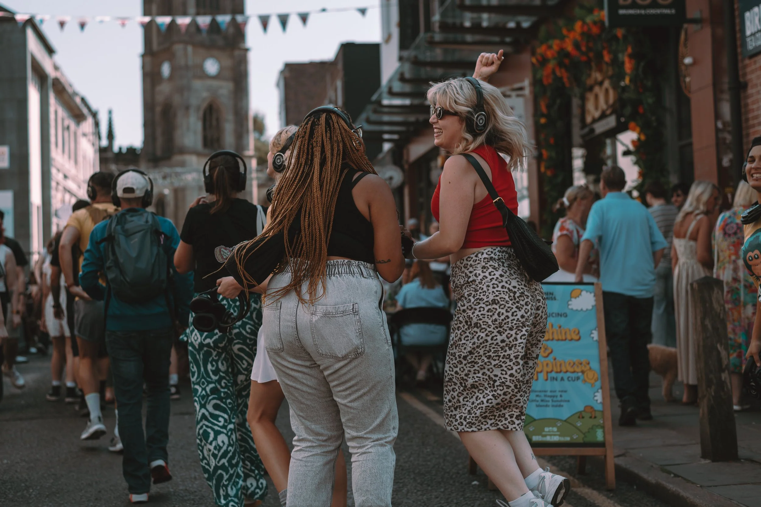 Two women dancing and smiling on a busy street with people walking in the background, wearing headphones.