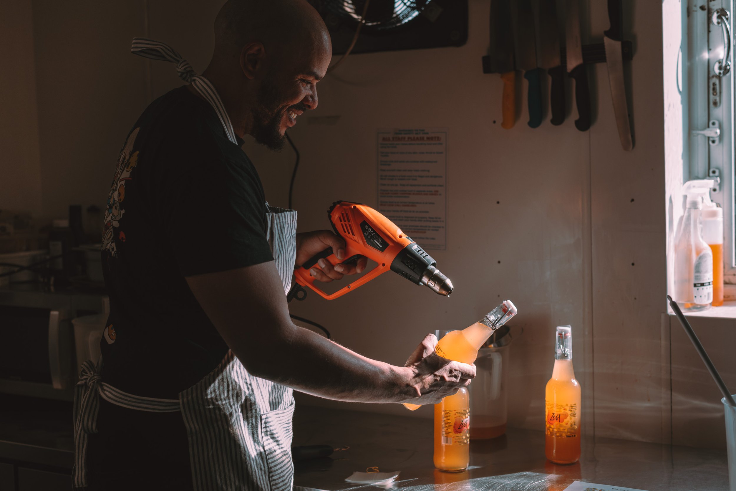 A man smiling while holding a bottle of orange-colored beverage and a heat gun in a kitchen. There are three identical bottles on the counter, with sunlight coming through a window.