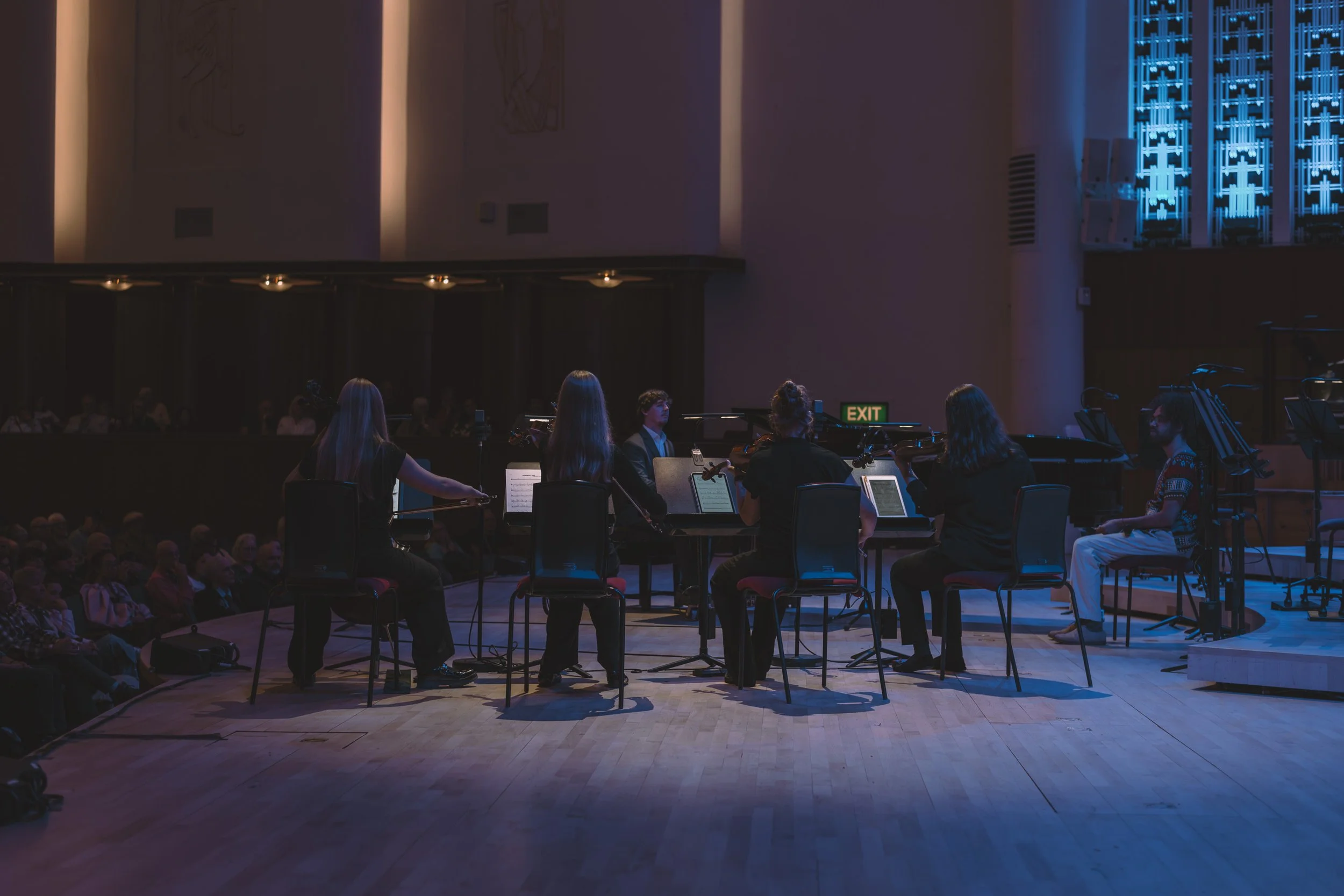 An indoor concert hall with a small ensemble of six musicians performing, seated in a semi-circle with music stands and instruments, audience seated in the background.