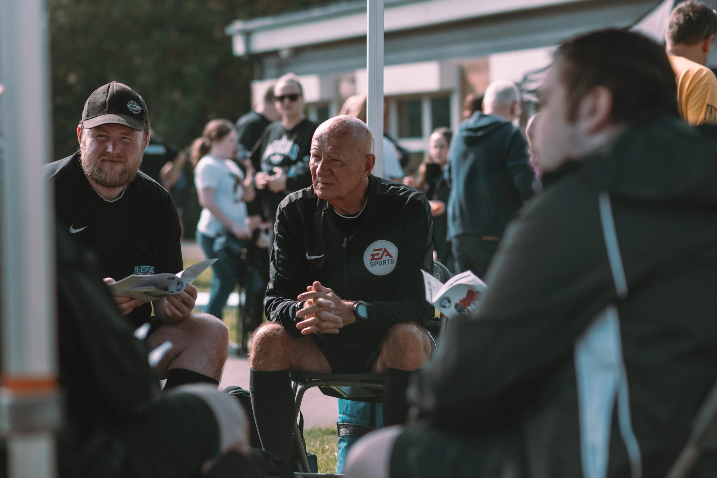 Group of men in black sportswear sitting and talking at an outdoor event, with a crowd of people in the background.