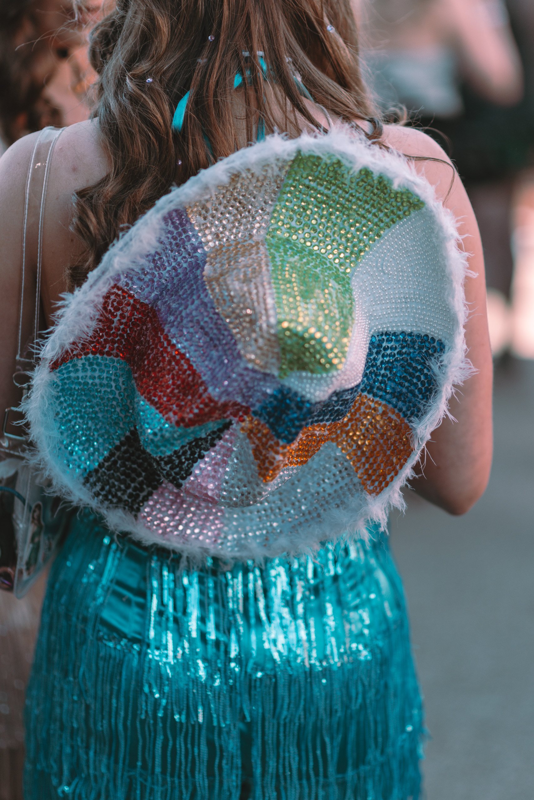 Person with long wavy hair wearing a colorful, oversized beaded hat with a fuzzy white brim, and a blue fringed skirt.