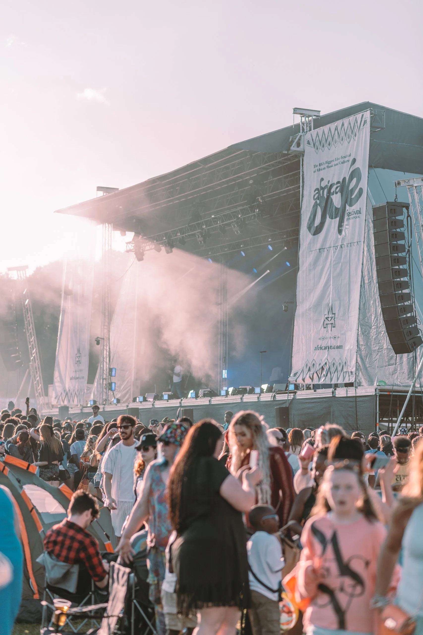 Crowd of people attending an outdoor music festival with a large stage, festival banners, and sunlight streaming in from the horizon.