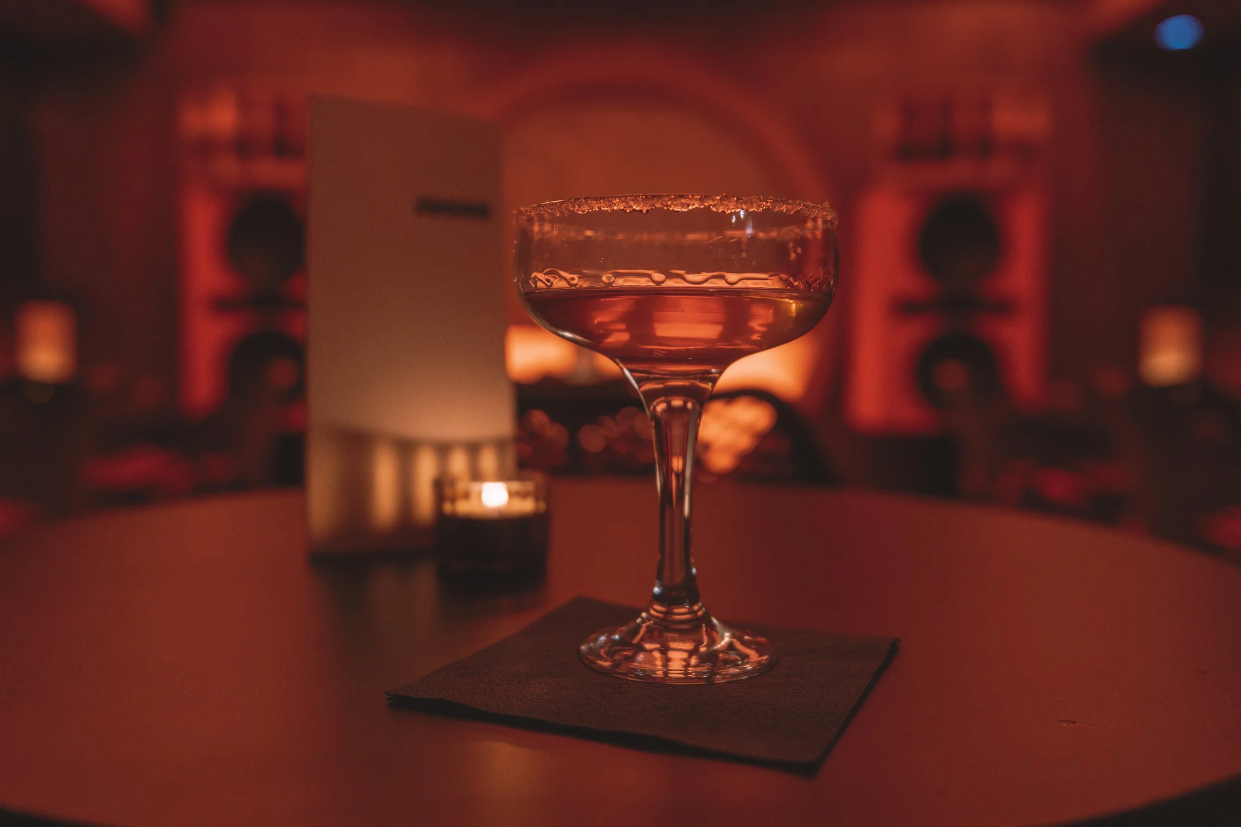 A cocktail glass with a salted rim filled with a reddish-orange drink, on a dark bar table with a black napkin, in a dimly lit bar or lounge with red lighting and blurred background.