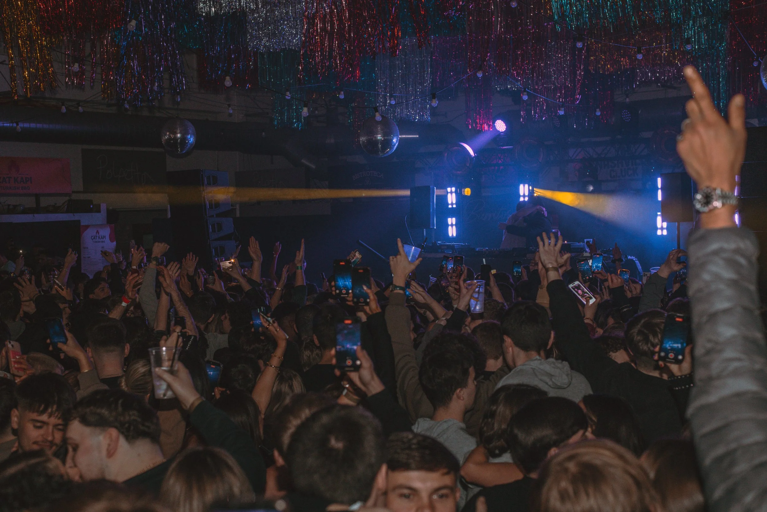 Crowd at a concert or party with colorful decorations and stage lighting, many people raising their hands and taking photos with smartphones.