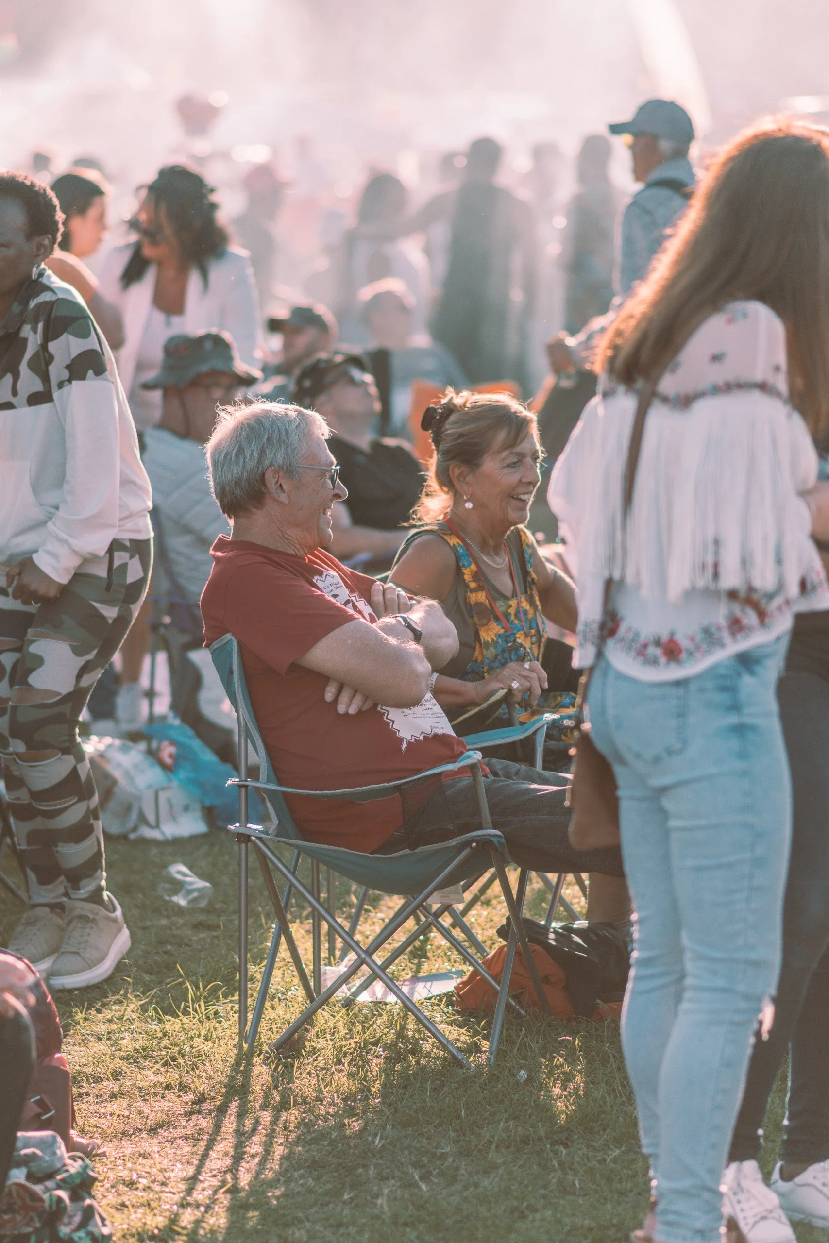 People gathering at an outdoor event during sunset, some sitting and others standing, engaging, with trees and horizon in the background.