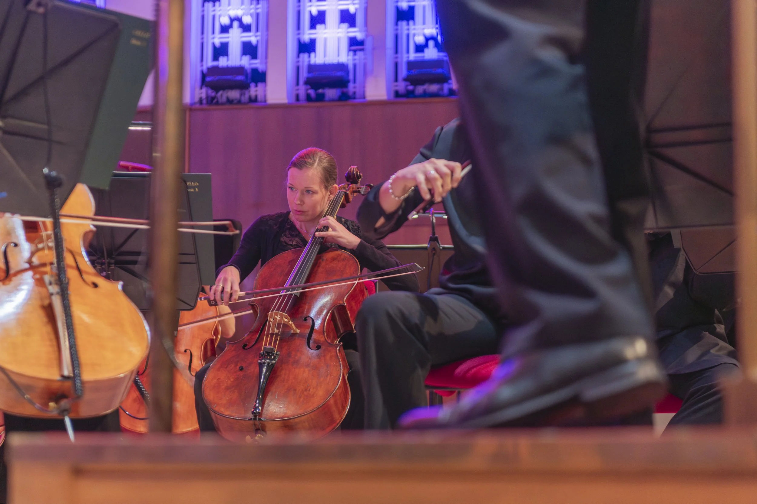 A woman playing a cello during an orchestra performance, with another musician partially visible in the foreground, in a concert hall with purple and blue lighting.
