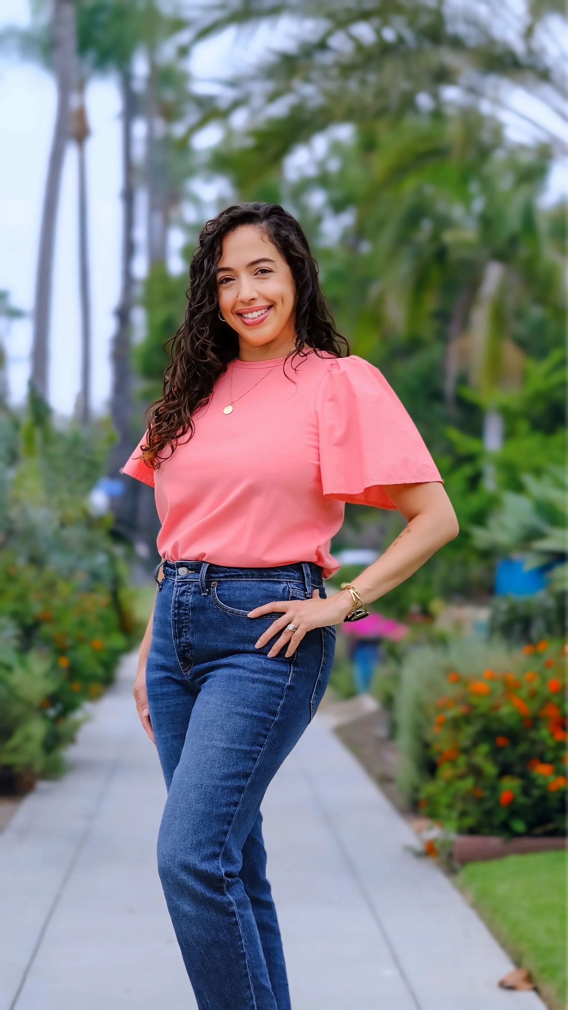 A woman with curly dark hair smiling and standing outdoors on a sidewalk, wearing a pink top and blue jeans, with a lush green background.