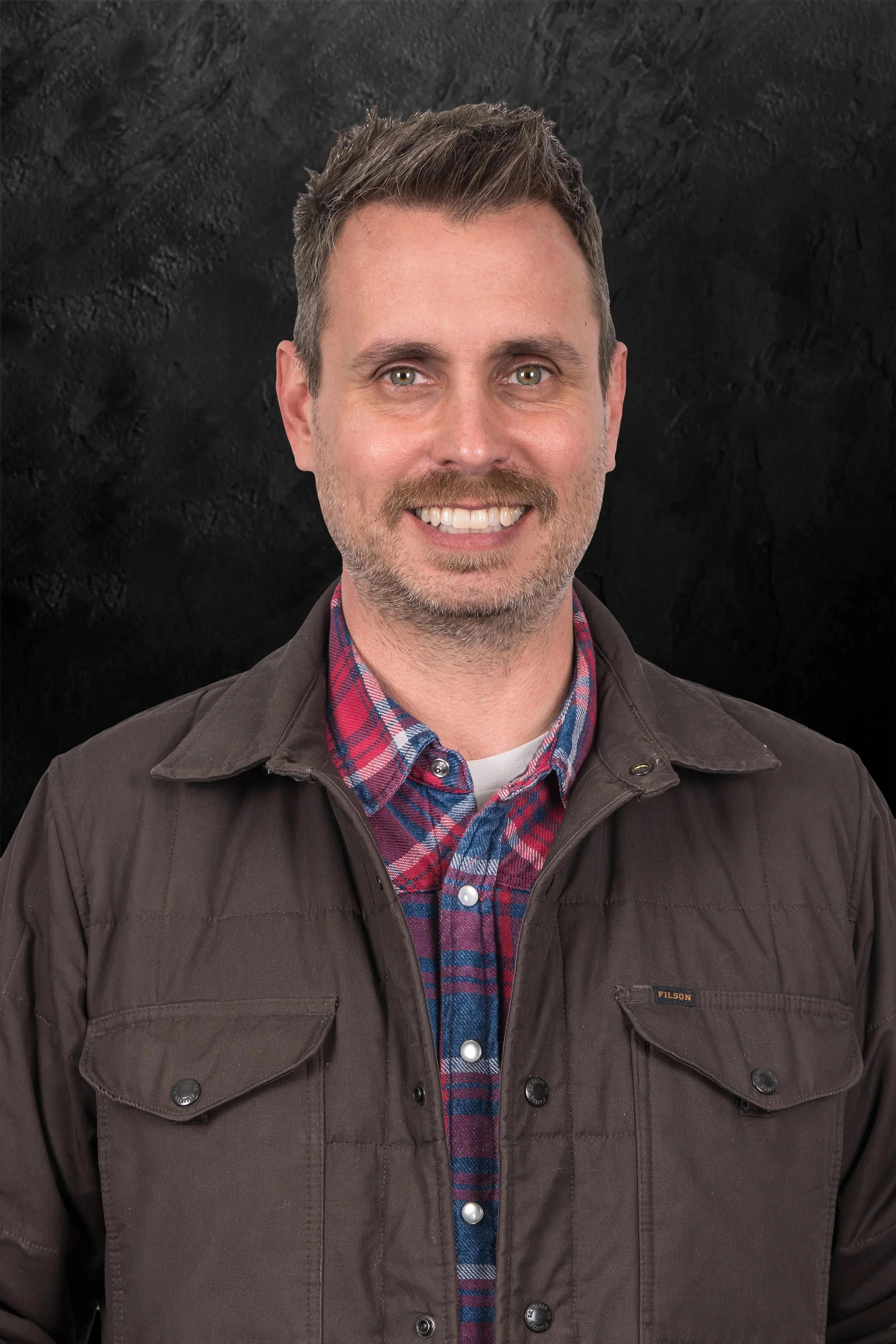 Smiling woman with long blonde hair wearing a green jacket against a dark background.