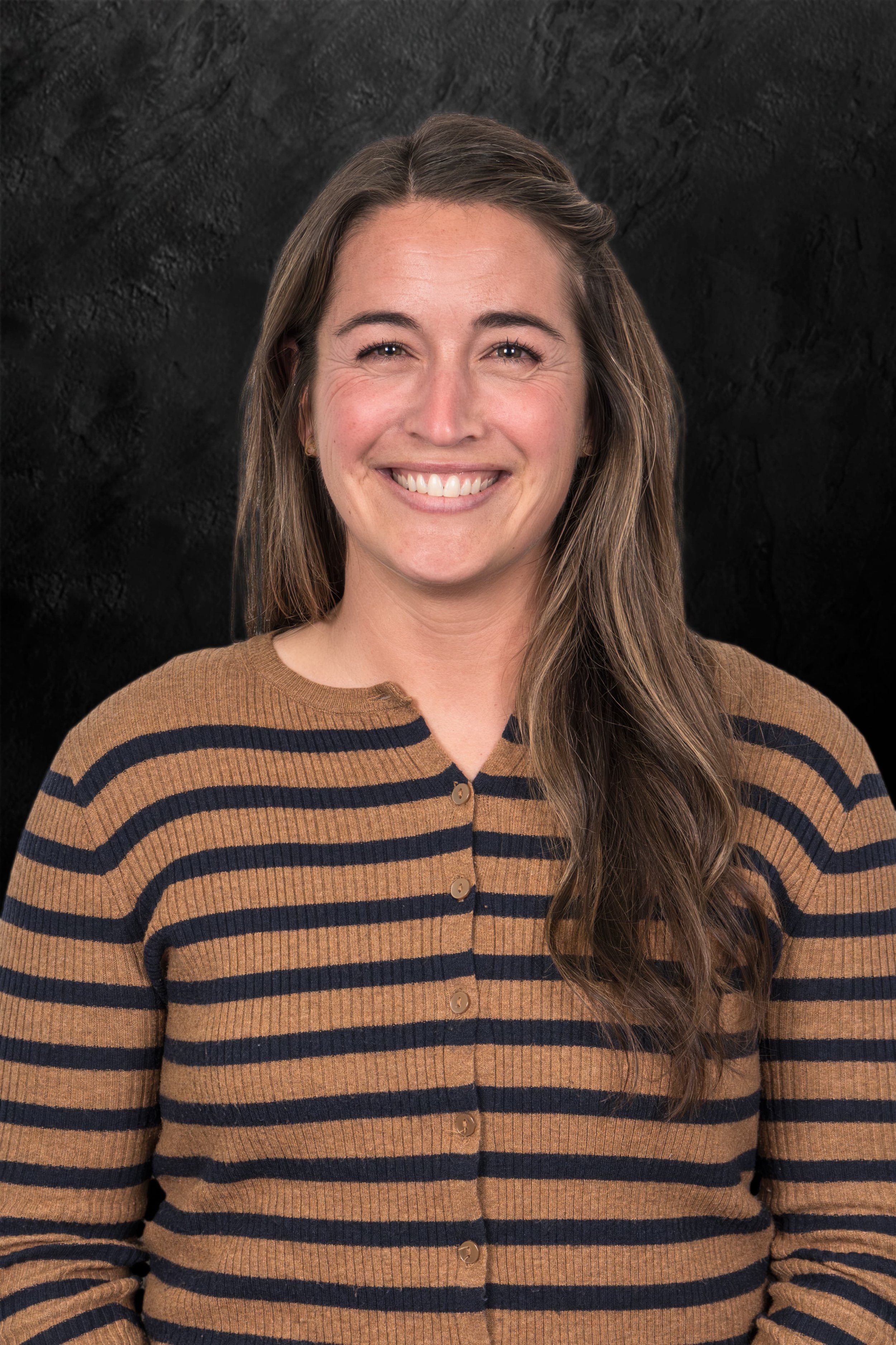 Portrait of a woman with long hair and hoop earrings, smiling, wearing a green shirt and dark jacket against a dark textured background.