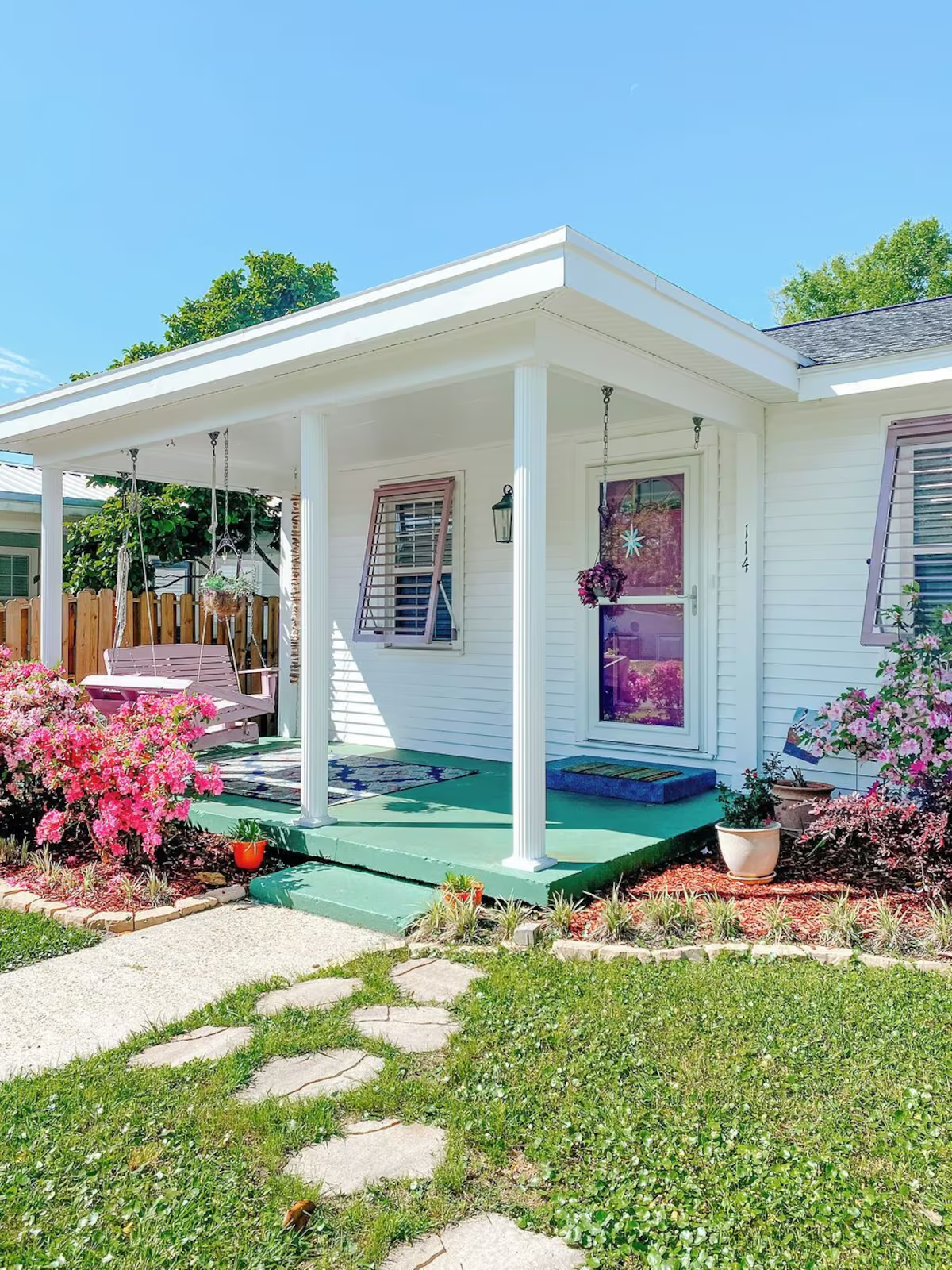 A front porch with purple swing, potted plants, pink flowering bushes, and a white house with a door and windows, under a bright blue sky.
