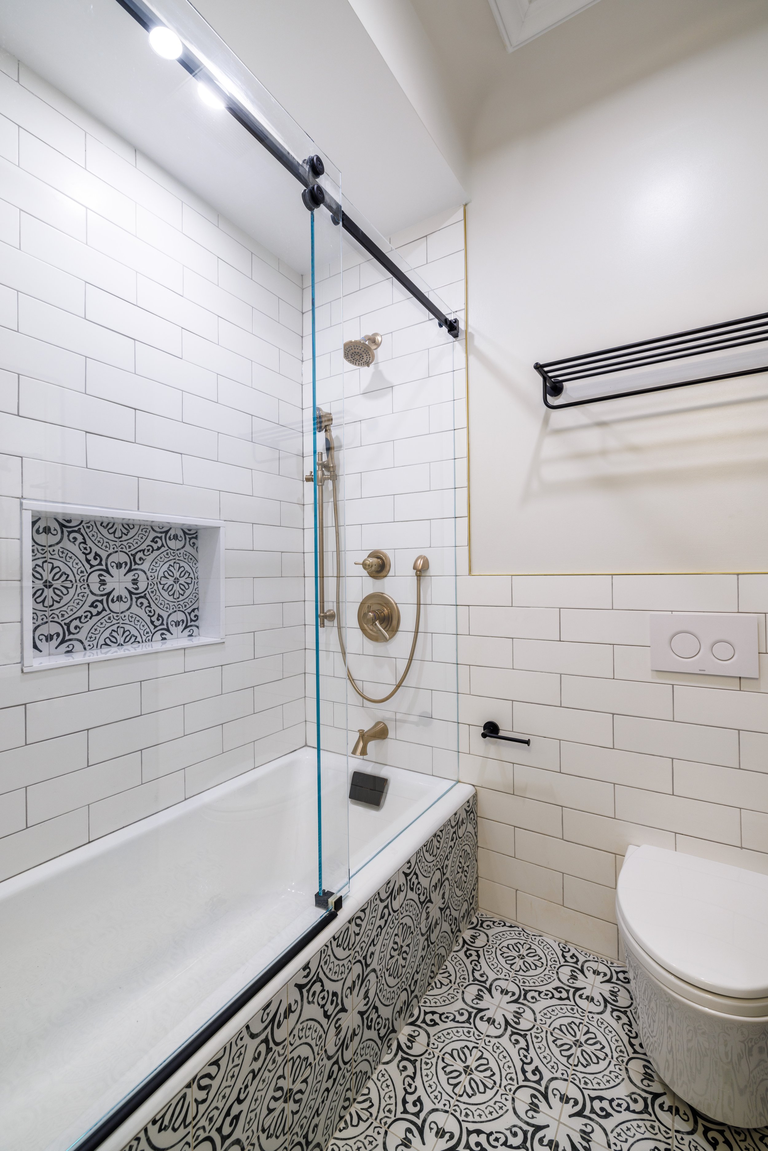 Modern bathroom with sliding glass shower door, white textured wall tiles, brass shower fixtures, bathtub, small window, and a toilet.