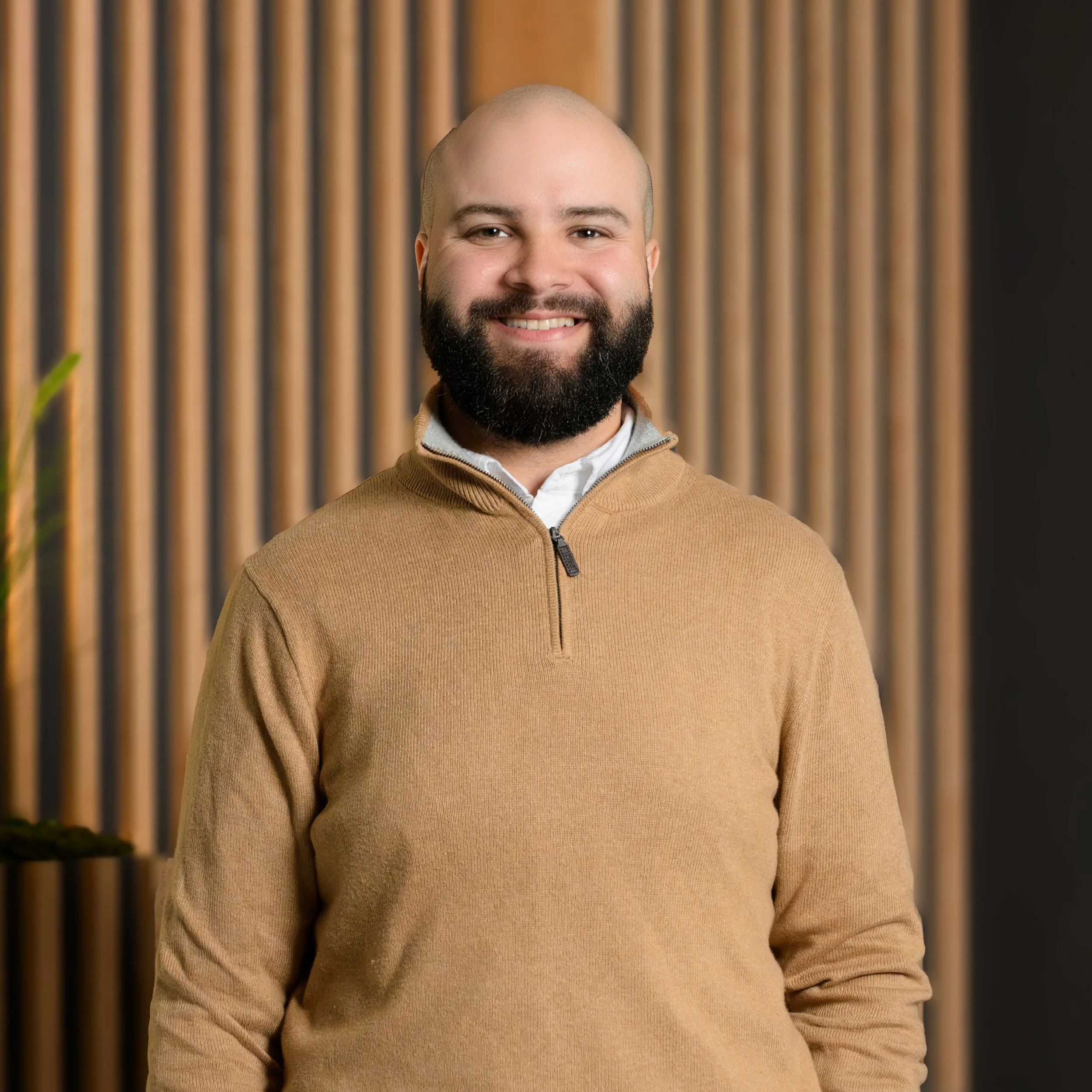 Smiling man with a beard in a beige sweater, standing in front of a wooden slat wall.