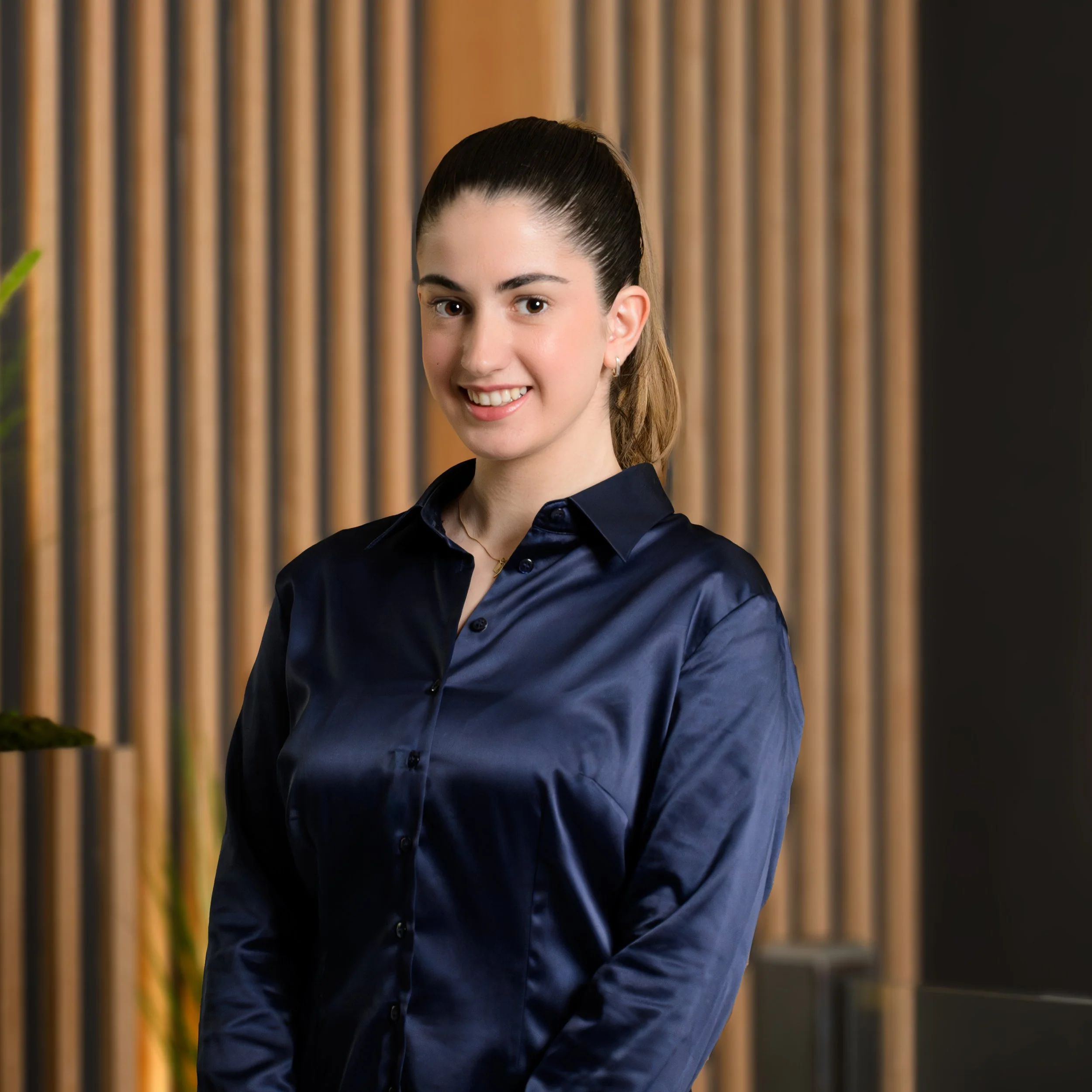 A woman in a dark blue blouse smiling, standing in front of a wooden panel background.