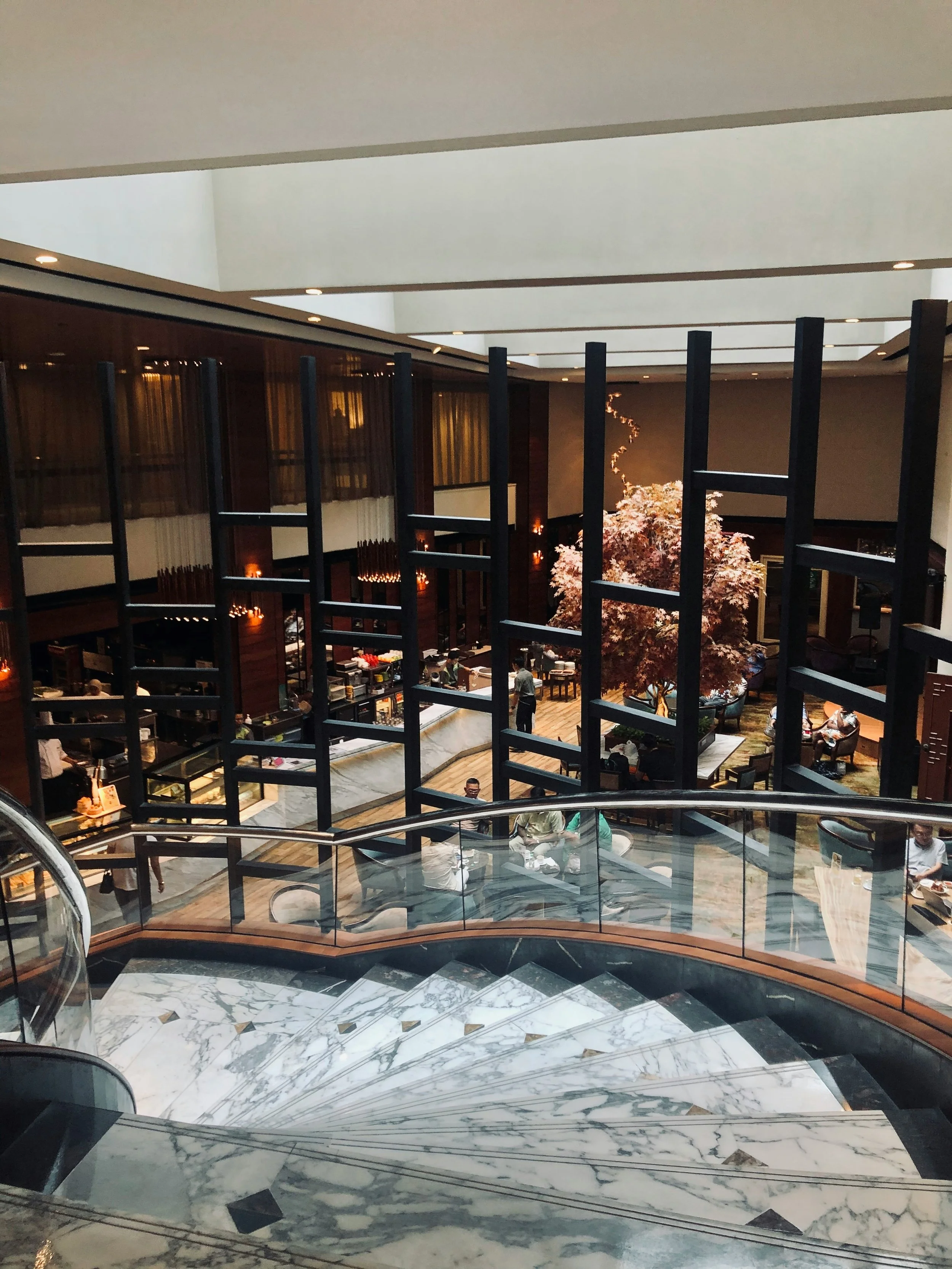 Modern interior with spiral marble staircase leading to a lower level with a restaurant. Features large decorative tree, seating area, and contemporary design elements.