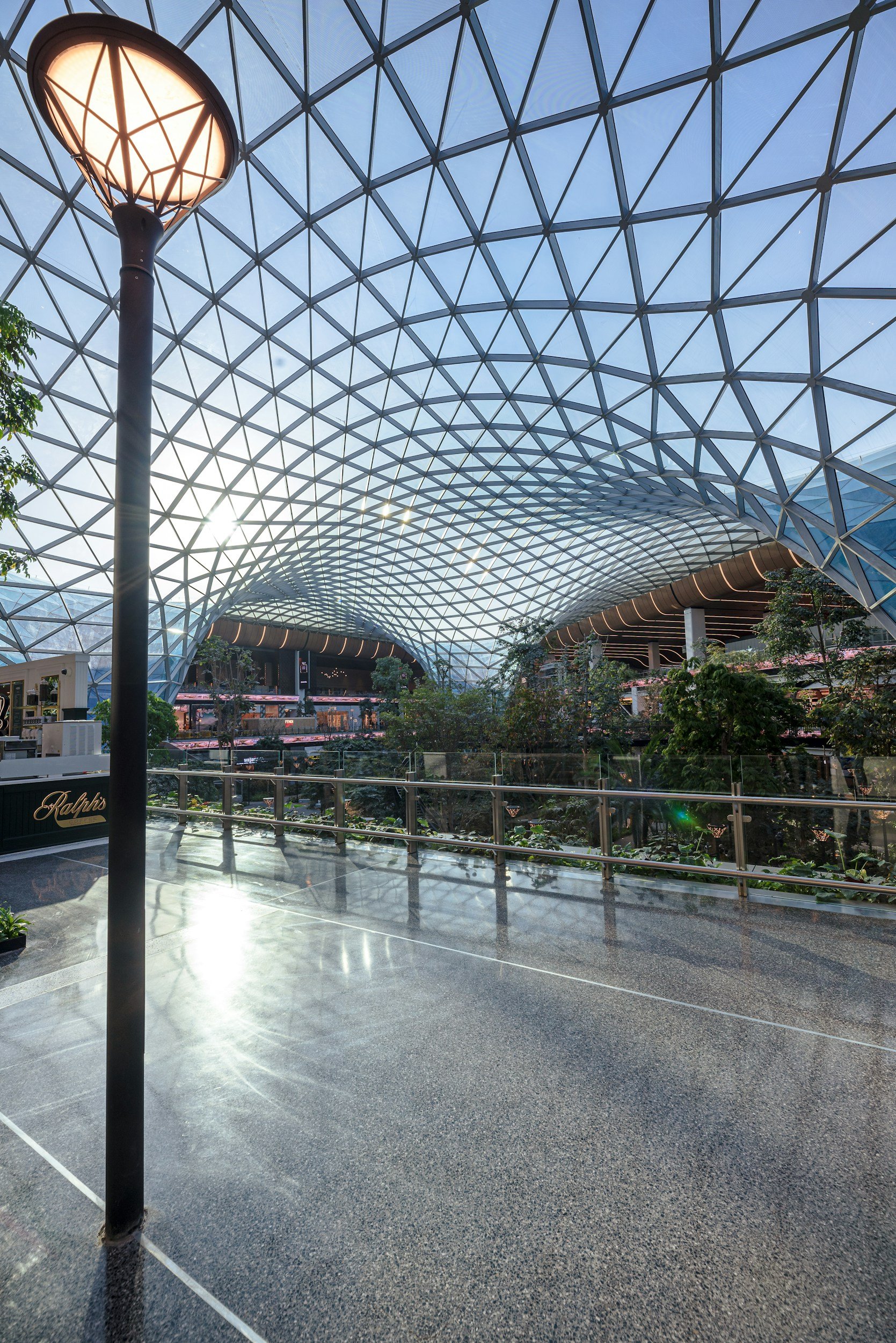 Interior of a modern building with a large glass dome ceiling, illuminated by sunlight, featuring greenery and glass railings.