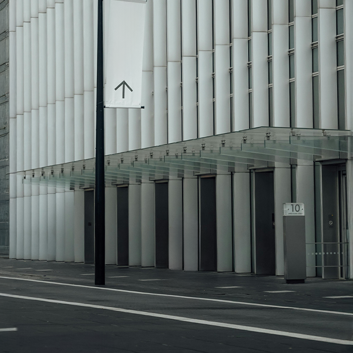 Modern building facade with glass awning and vertical panels, alongside a street with a white directional sign and a pole.