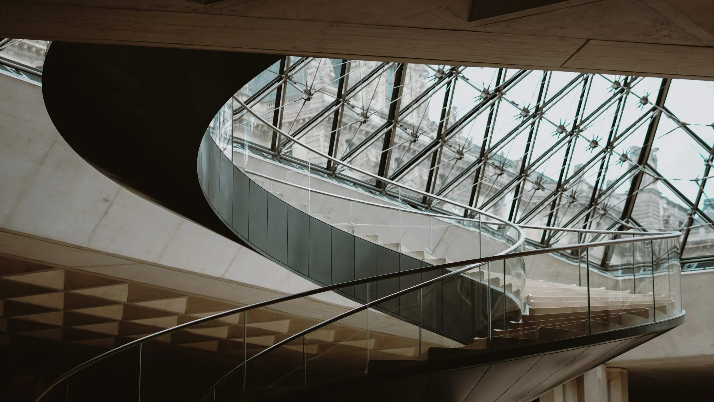 Spiral staircase with glass railings and a modern architectural background featuring a glass-roofed structure.