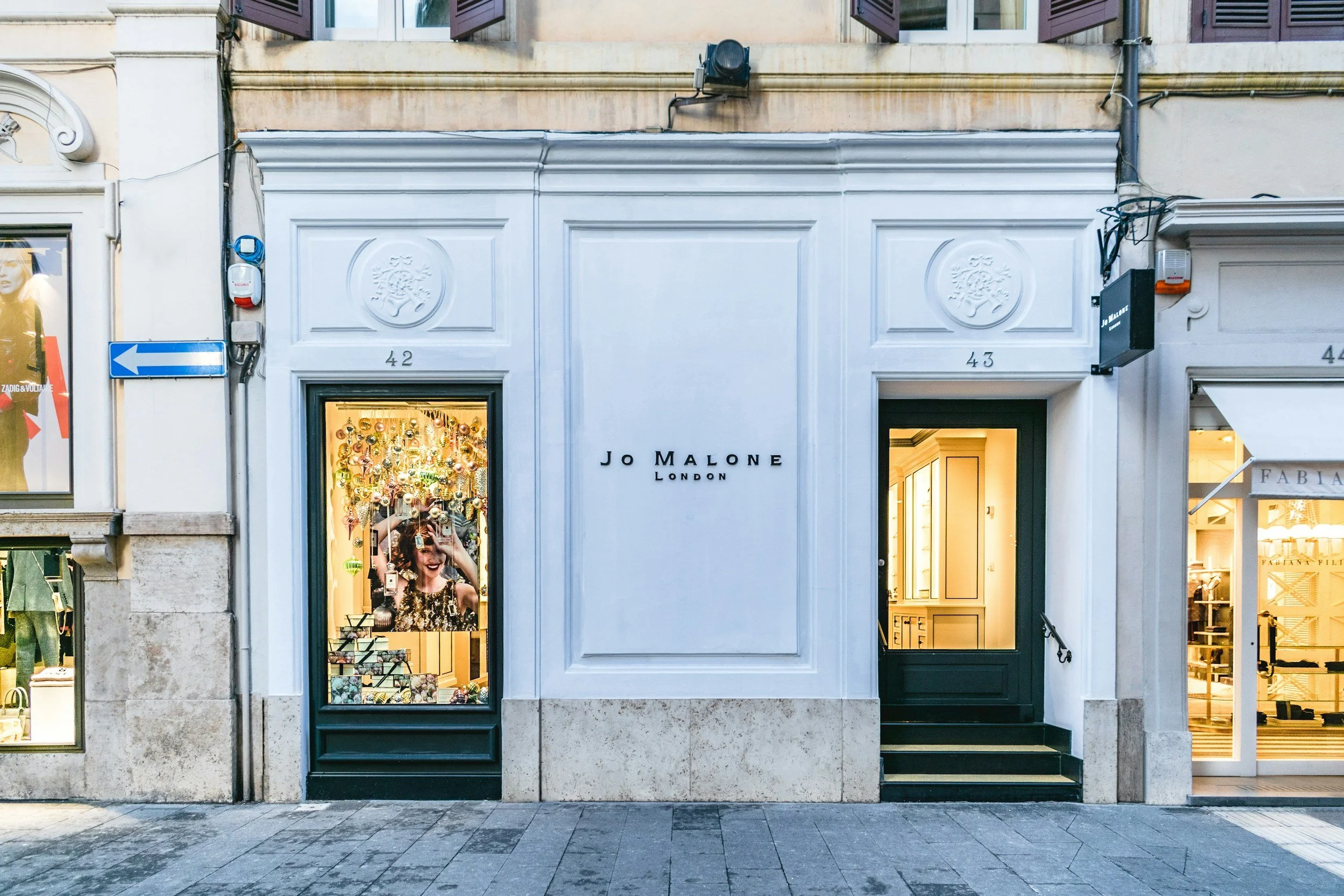 Exterior of a Jo Malone London store with a white facade and two black-framed windows. The left window displays decorative items and a person, while the right shows an illuminated interior. Building numbers 42 and 43 are visible.