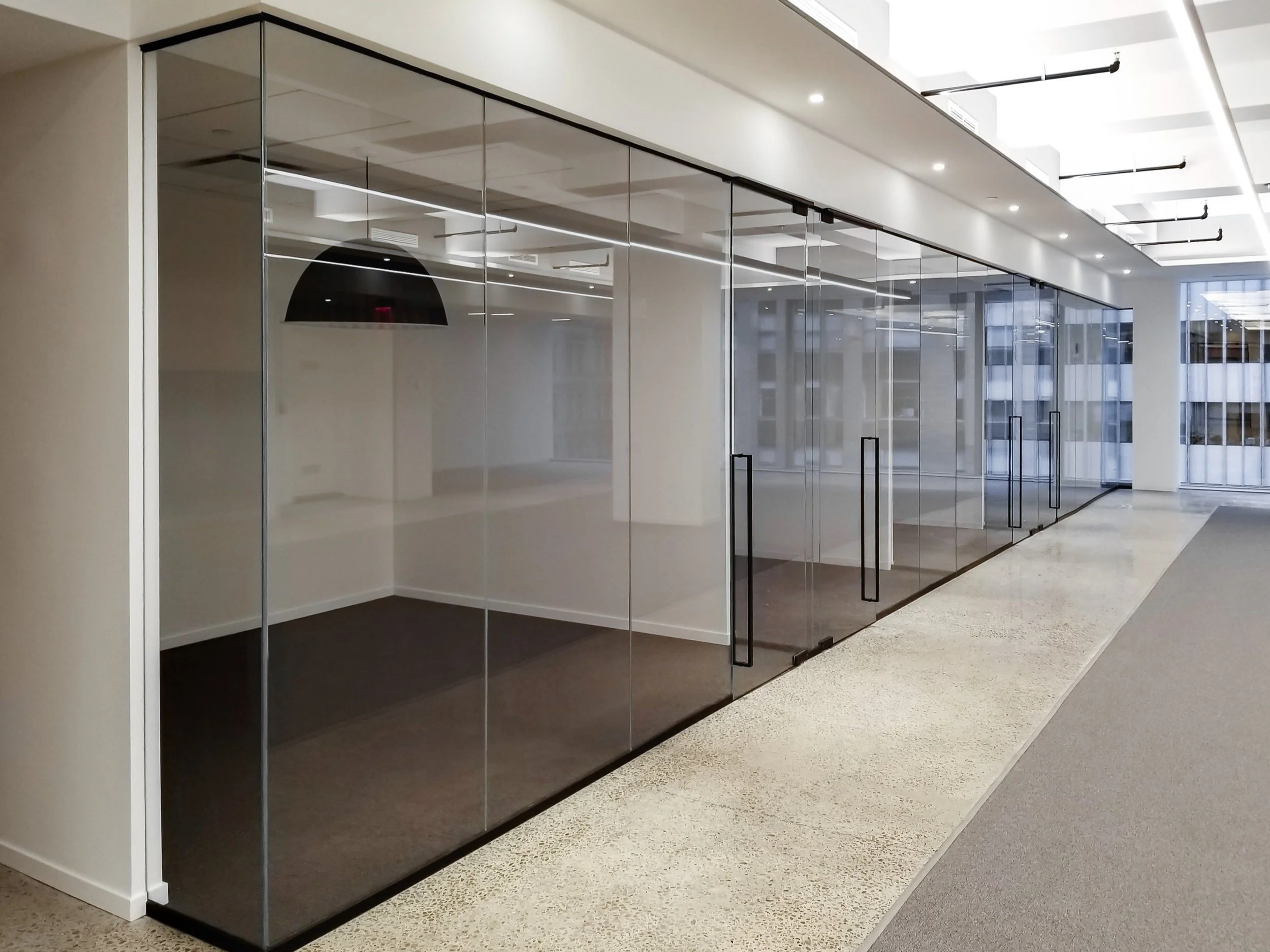 Empty office space with glass walls and doors, modern ceiling lights, and large windows overlooking city buildings.