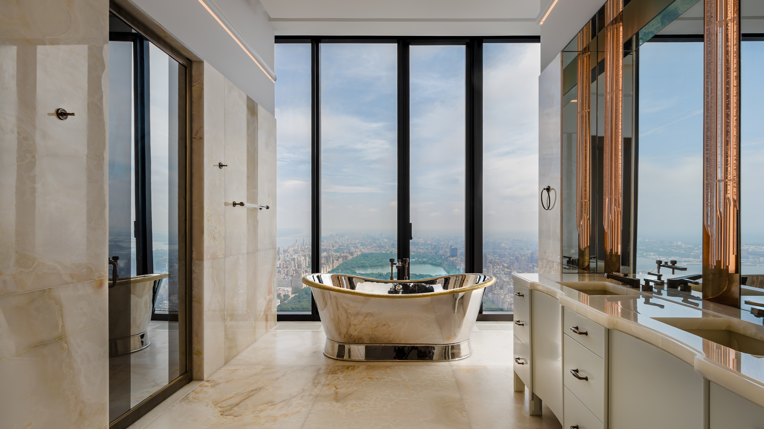 Luxurious bathroom with a gold bathtub, large floor-to-ceiling windows showing a city view with a park, marble walls and flooring, a double vanity with black faucets, and modern lighting fixtures.