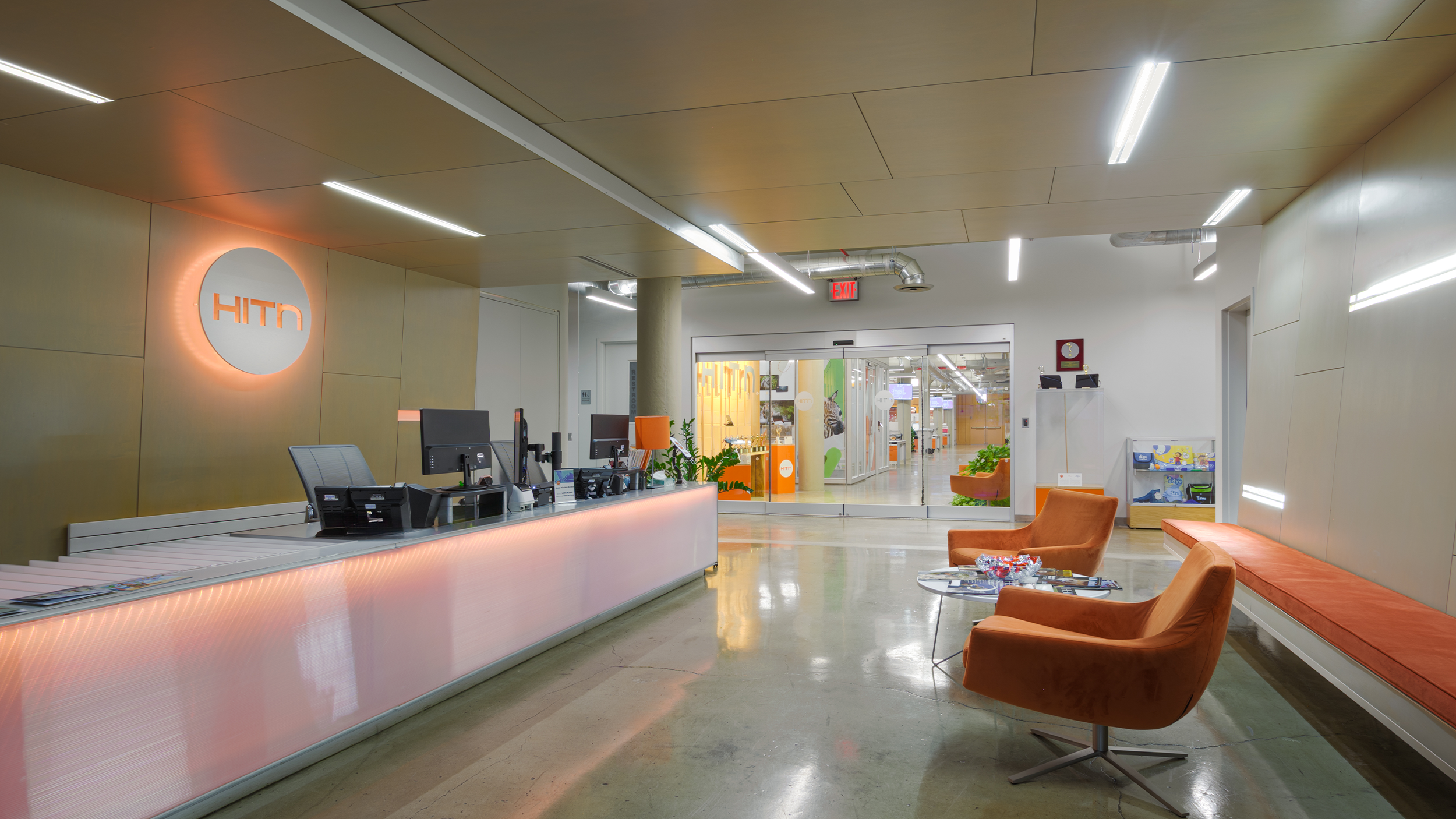 Modern office reception area with a curved white front desk, orange and brown chairs, plants, and glass doors leading to an open workspace, illuminated by ceiling lights.