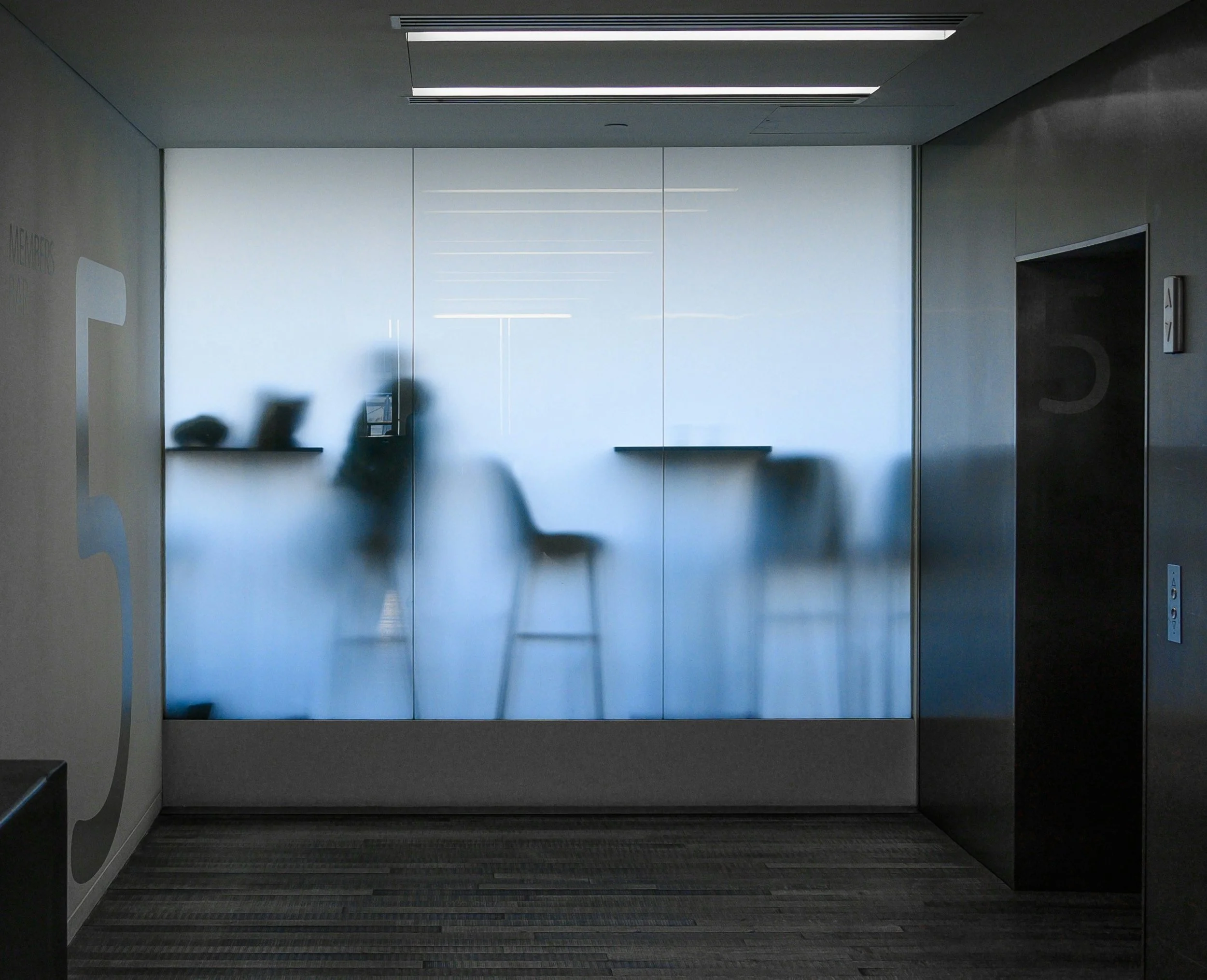 Blurred silhouettes of people and furniture behind frosted glass in a modern office setting.
