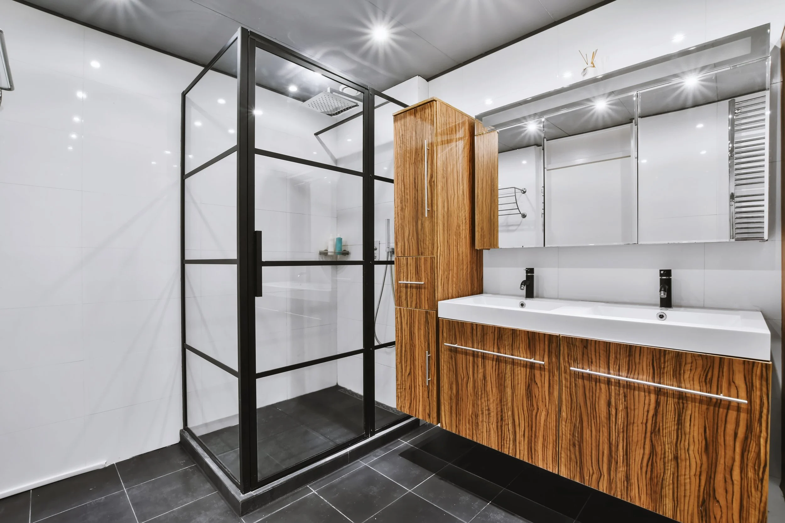 Modern bathroom with a glass shower enclosure, wooden cabinets, double sink, and tiled floor.