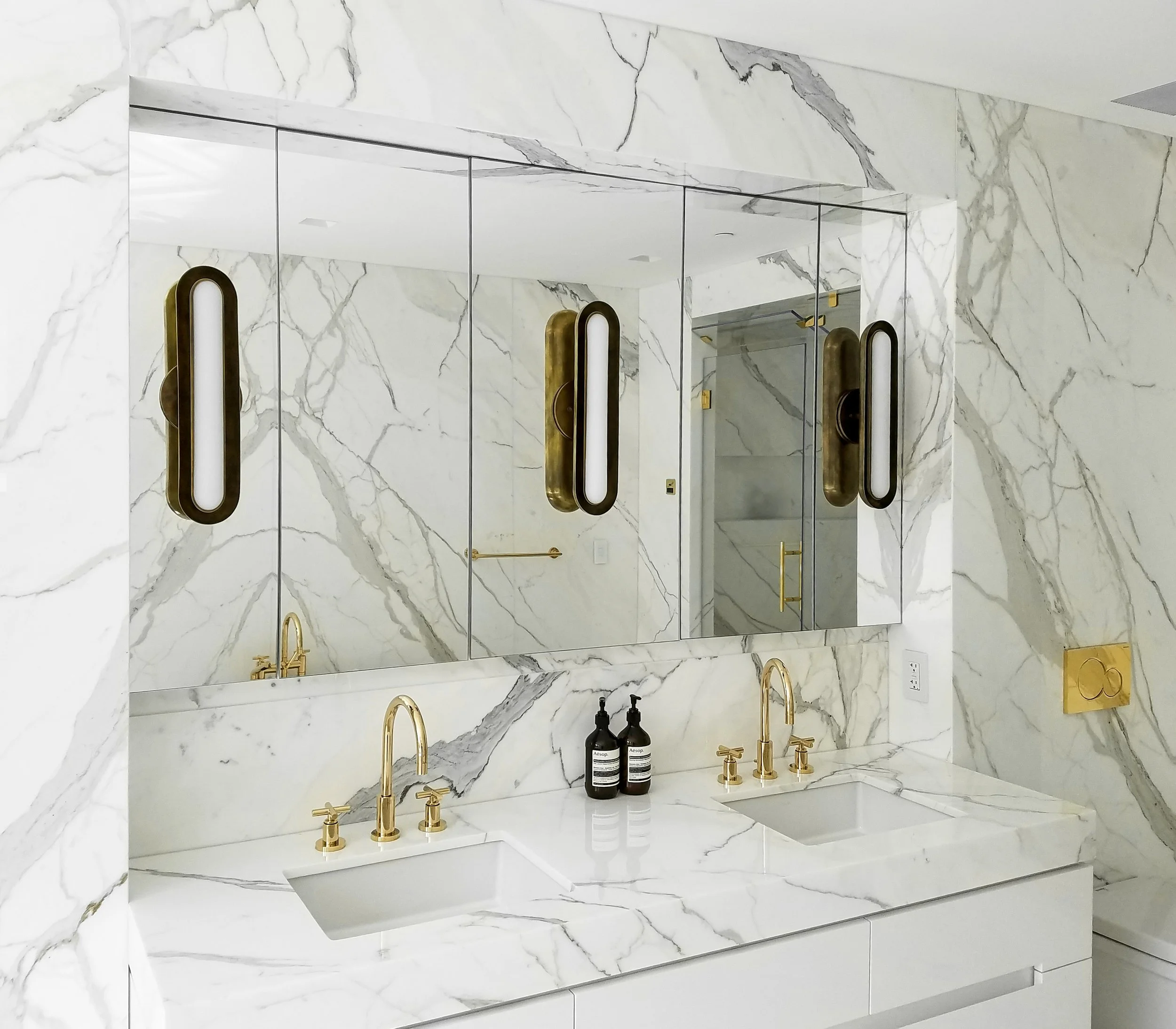 Modern bathroom with marble walls and countertops, featuring two sinks with gold faucets, mirrored cabinets, and two wall lights.